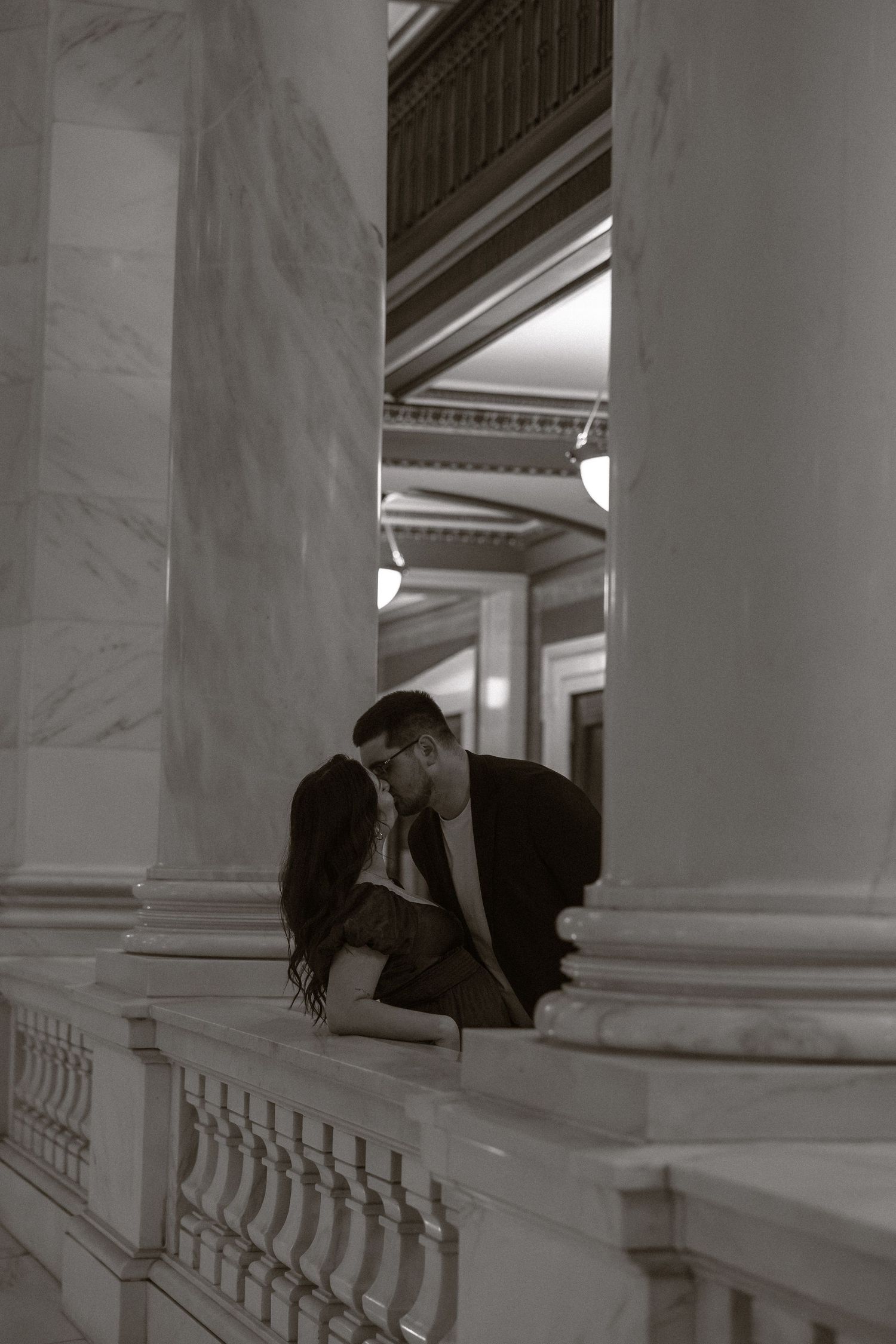A romantic black and white photograph of a couple sharing an intimate moment between elegant marble columns.
