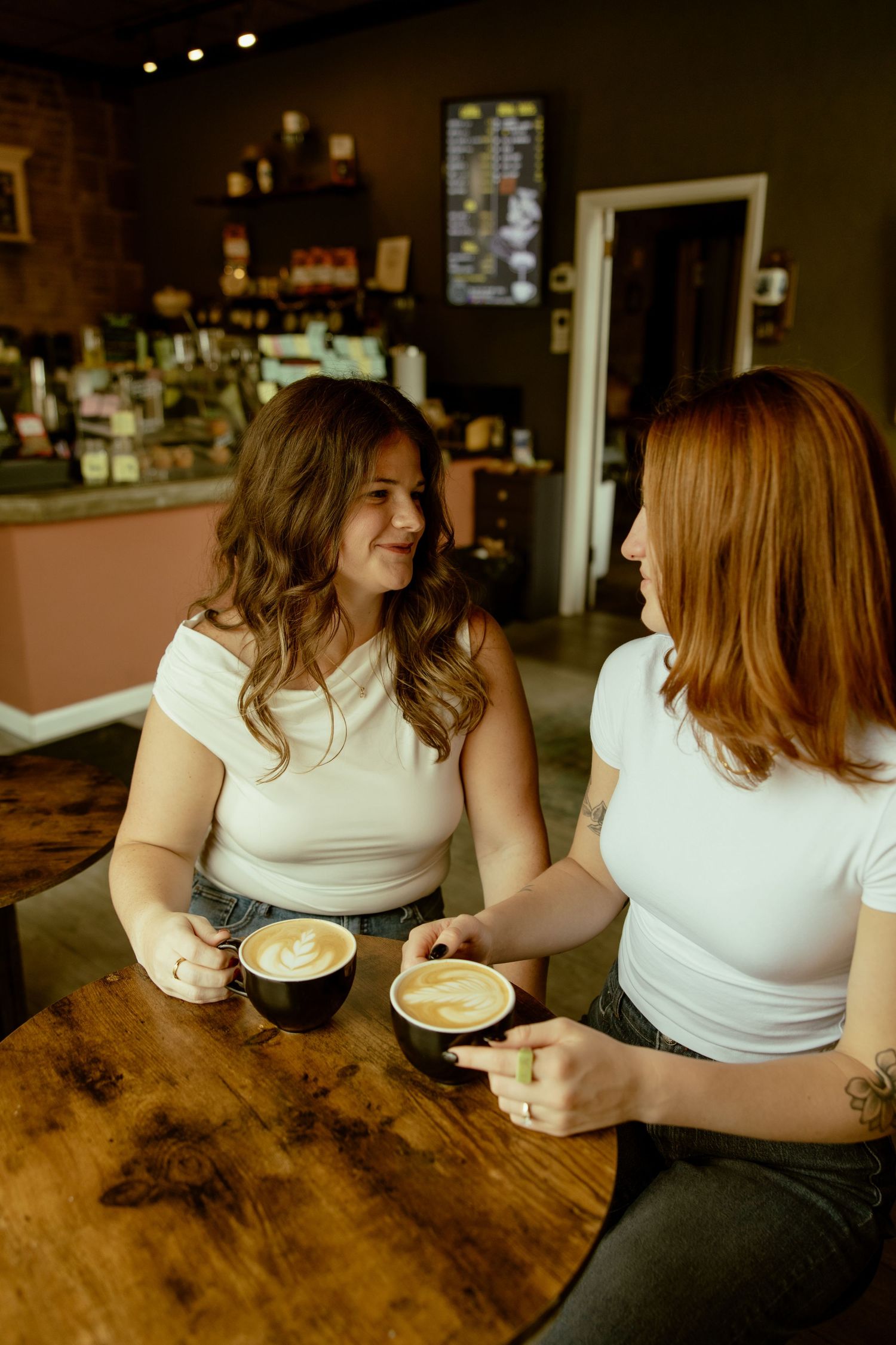 Two friends enjoying coffee with latte art while chatting at a rustic wooden bar counter in a cozy cafe.