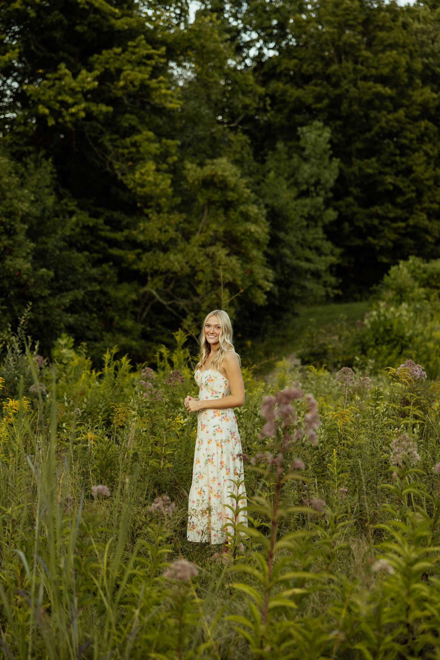 A person in a floral dress stands among wildflowers and tall grass in a sunlit meadow with trees in the background.