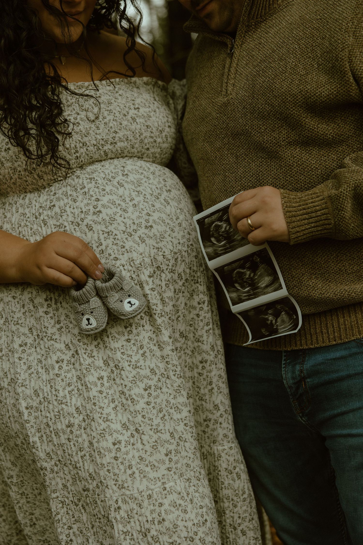 Person wearing brown sweater and jeans holds ultrasound photos while sitting on speckled carpet.