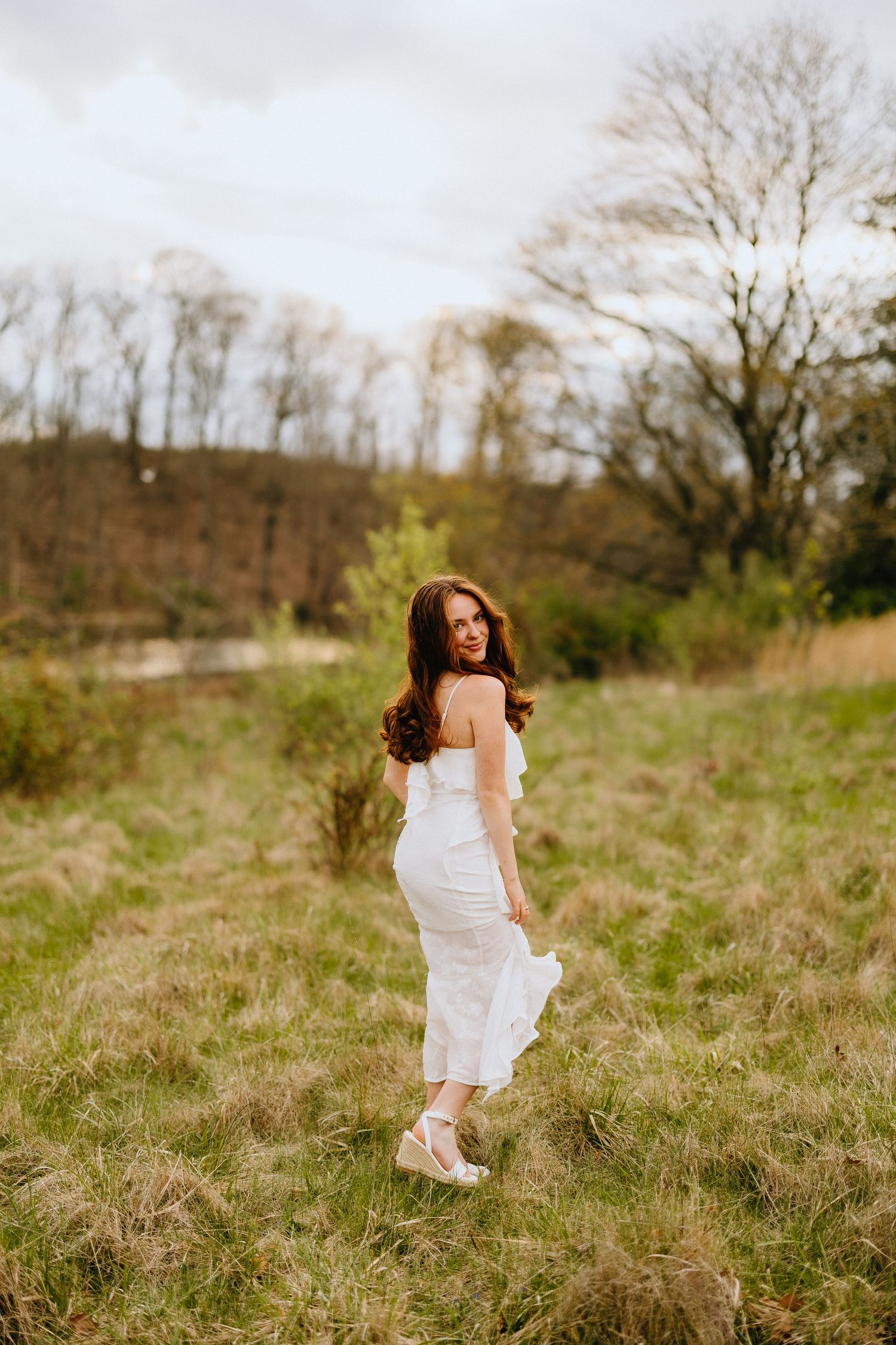 A graduate in a white dress poses in a sunlit meadow with trees in the background.
