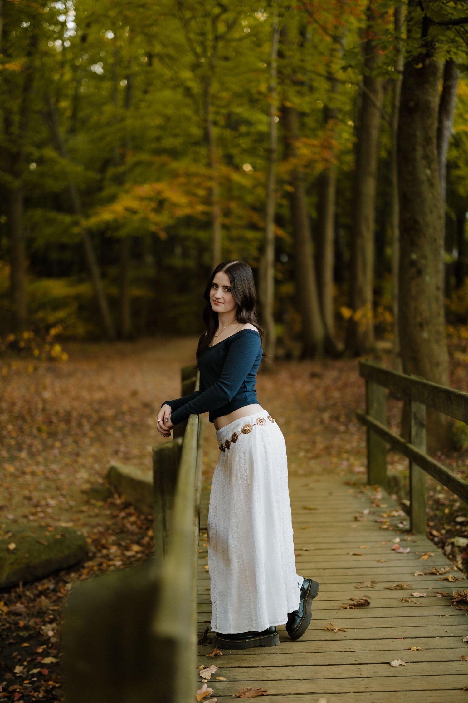 Figure in navy top and white skirt stands on wooden boardwalk in autumn forest.