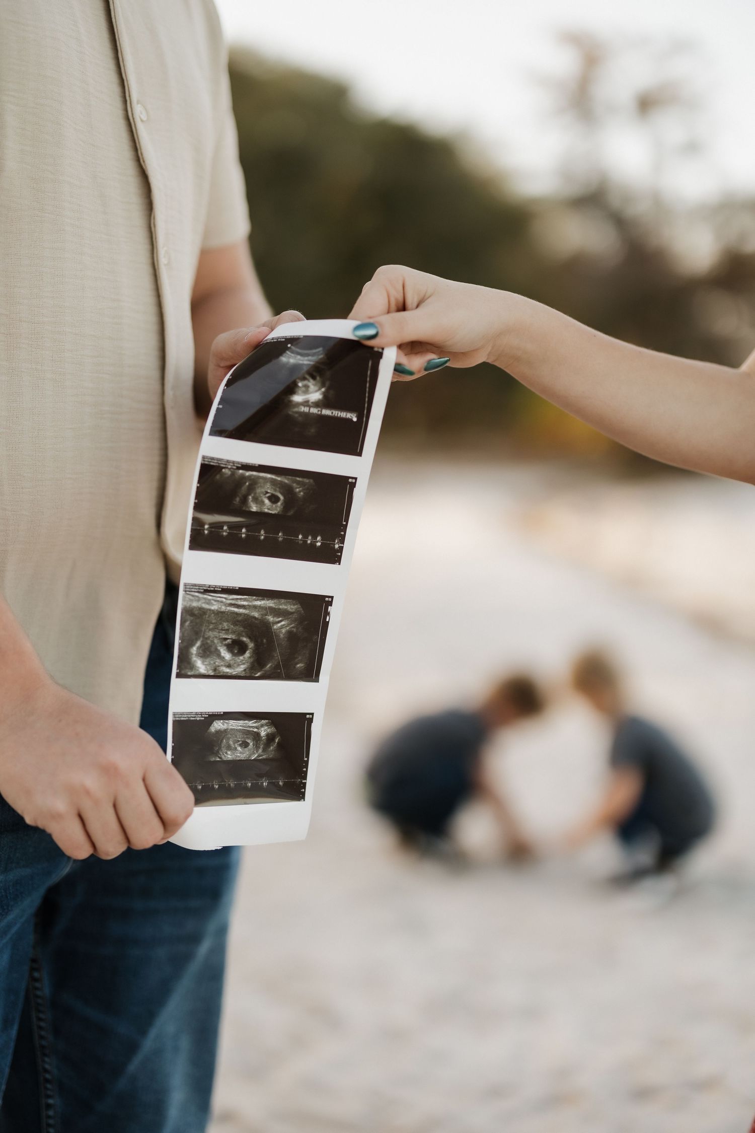 Close-up of hands holding ultrasound photos with blurred couple in background.