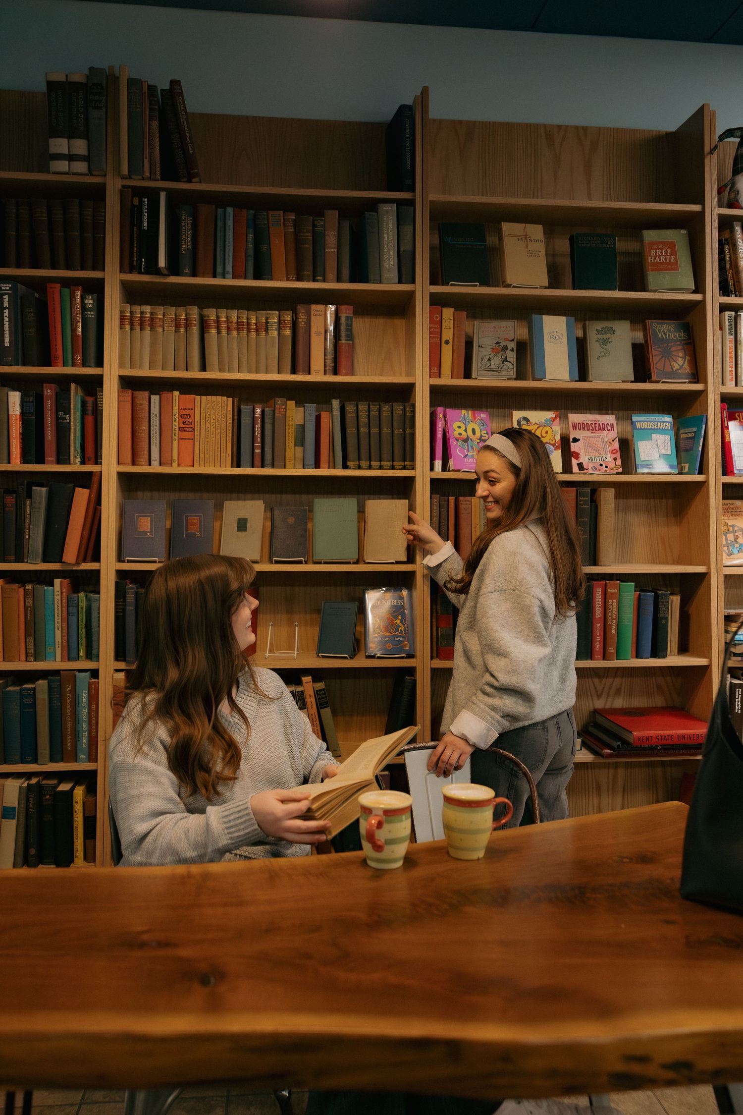 Friends enjoying coffee in a library with wooden table and floor-to-ceiling bookshelves.