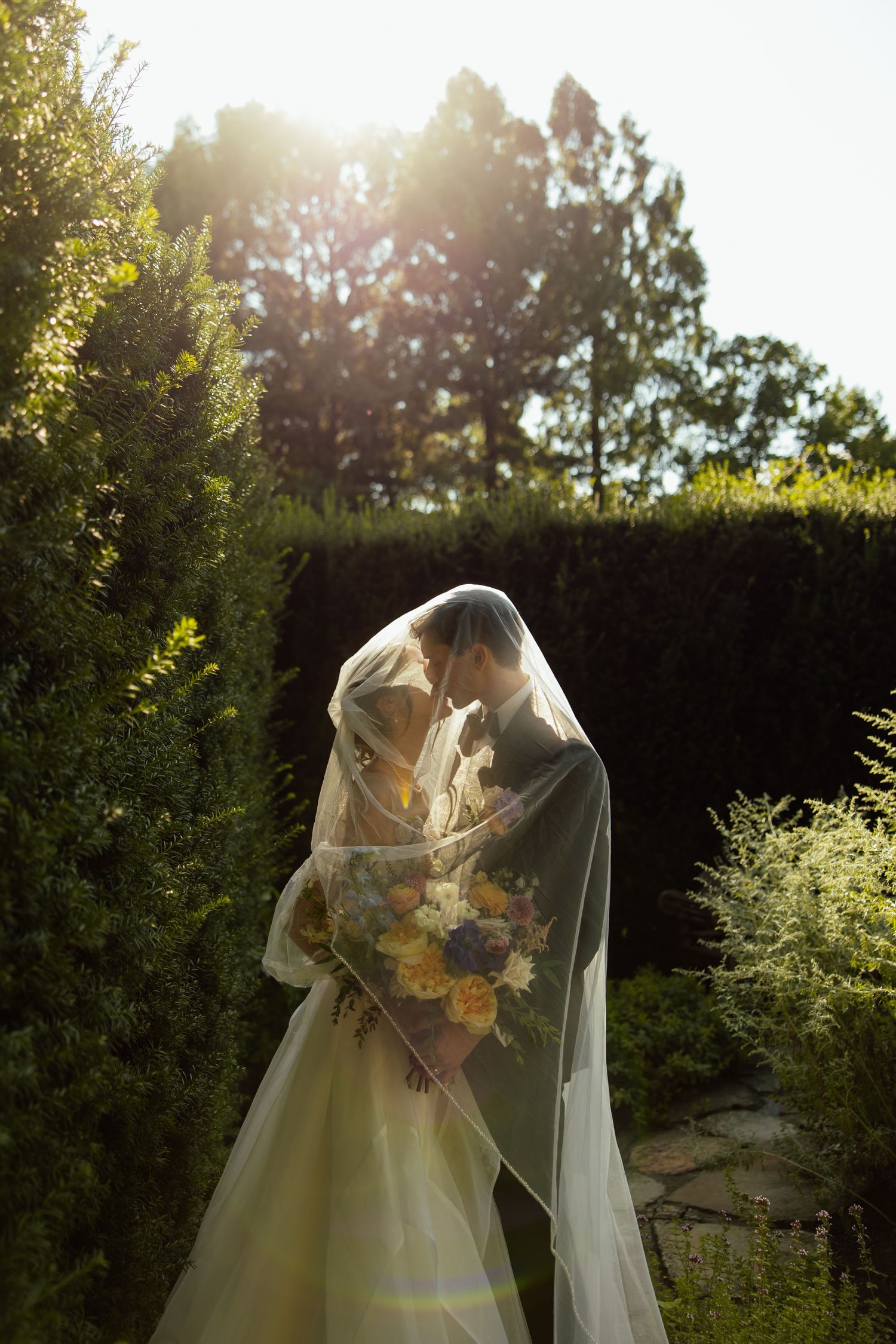 A bride in a flowing white dress and veil holds a sunflower bouquet in a sunlit garden with greenery.