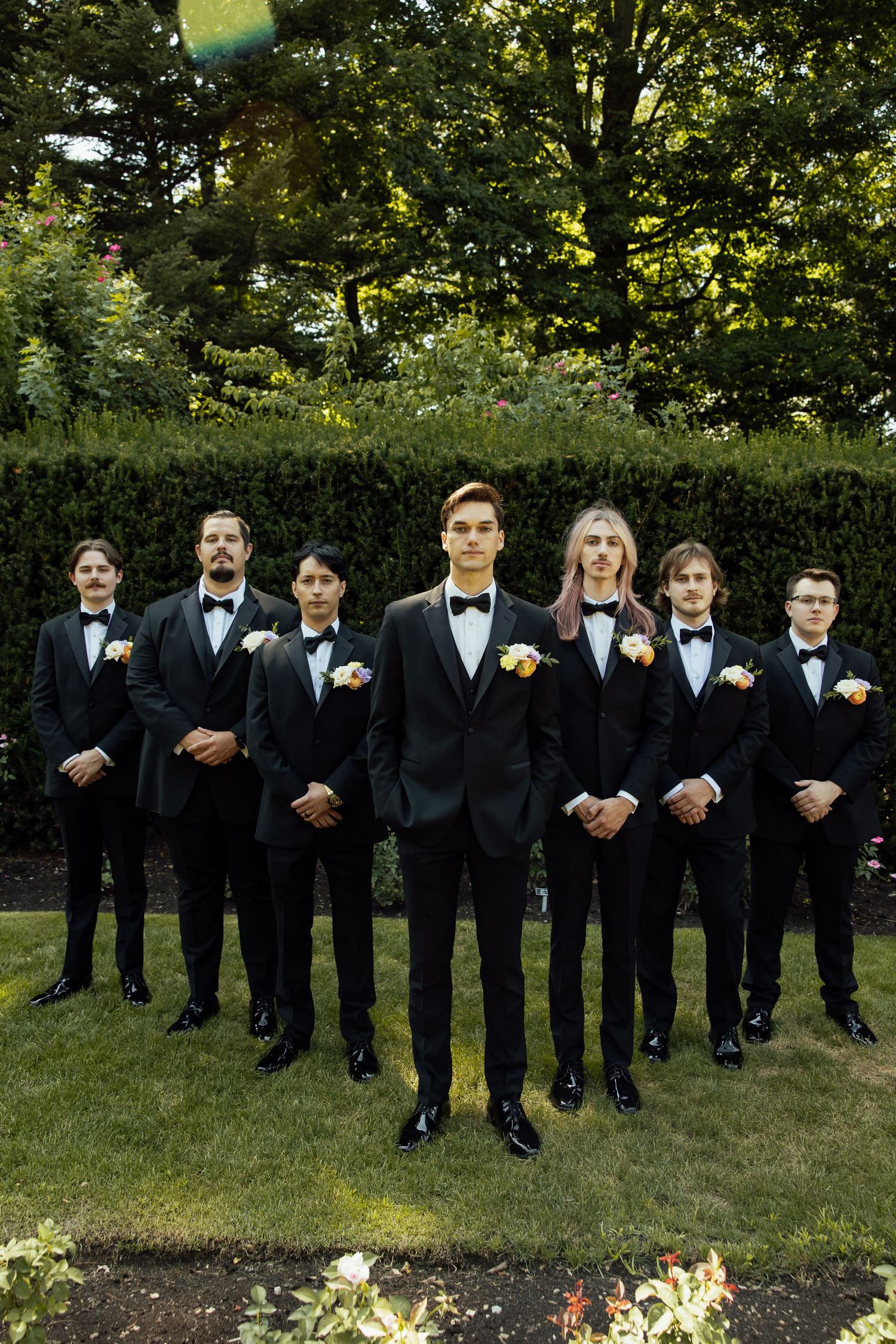 A wedding party of groomsmen in black tuxedos and bowties stand in a line outdoors on a sunny day.
