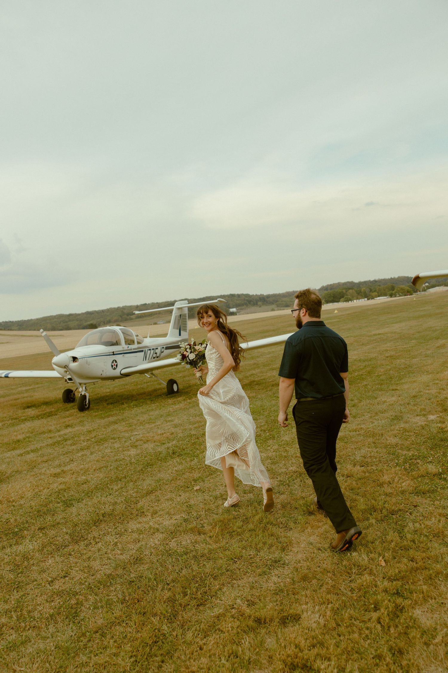 A wedding couple walks across an airfield with small planes in the background on an overcast day.