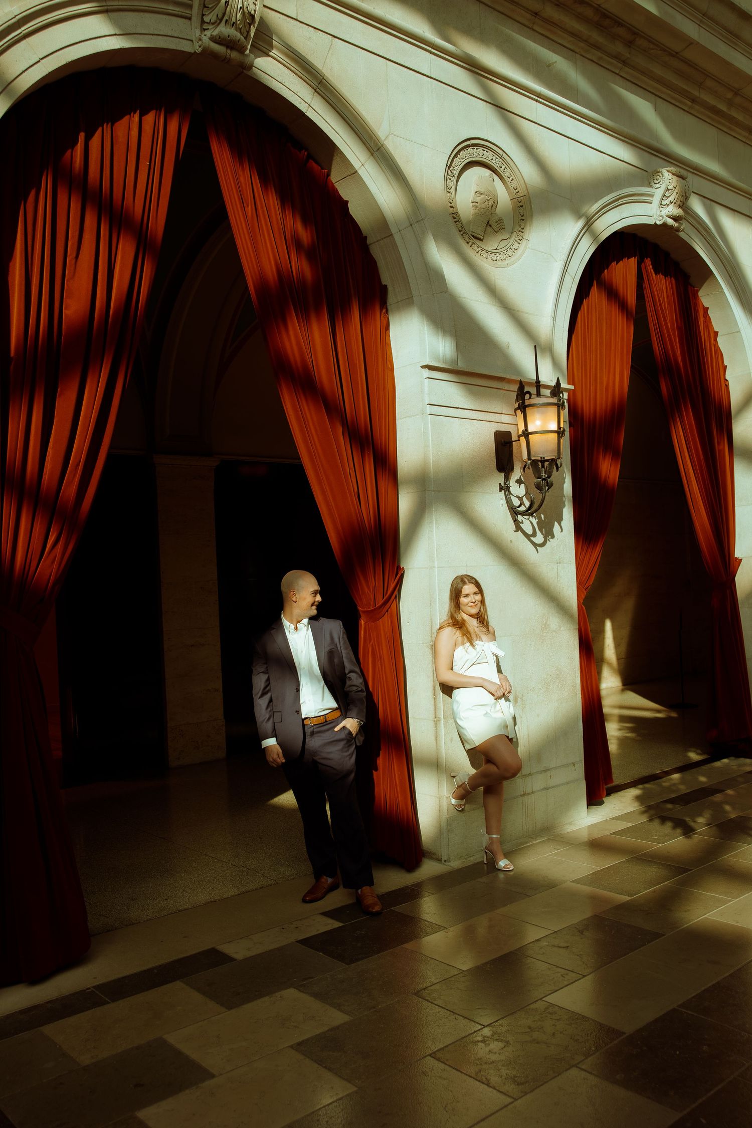 Dramatic lighting illuminates figures in formal attire against dark red curtains in an elegant hallway.