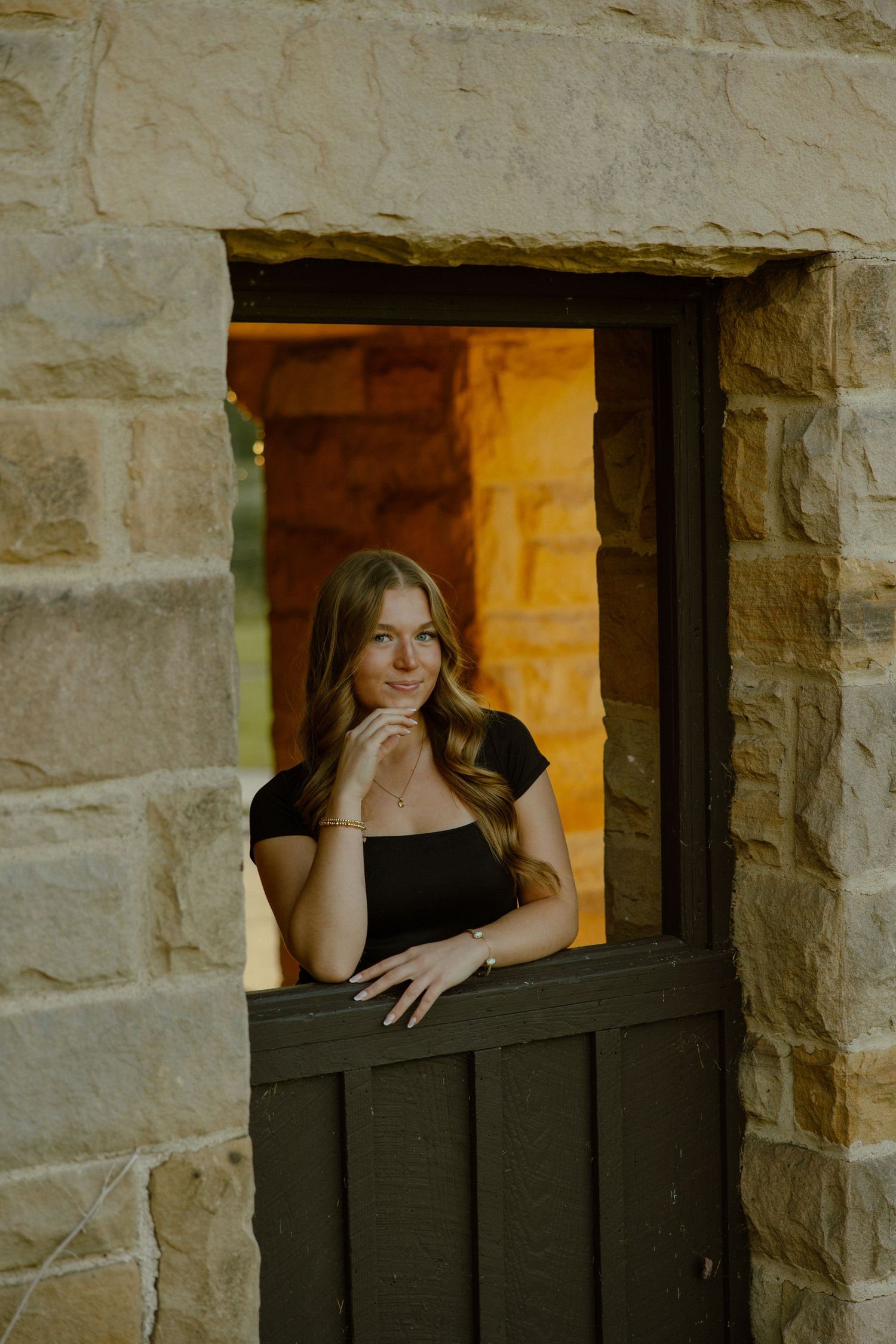 A rustic stone wall window frames a figure leaning on a dark wooden ledge in warm lighting.