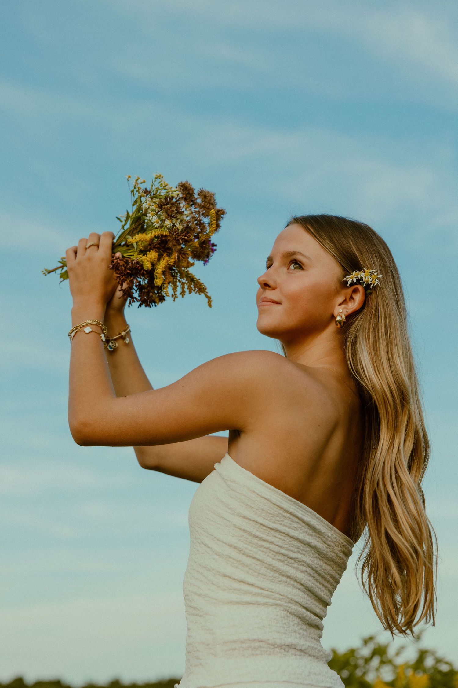 A person in a white strapless dress holds up dried flowers against a blue sky background in a series of artistic poses.