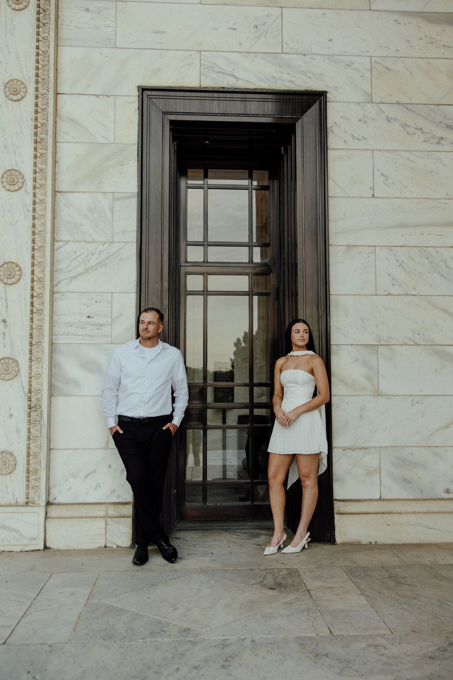 A couple poses together against a white marble building with dark doorways during a photo shoot.