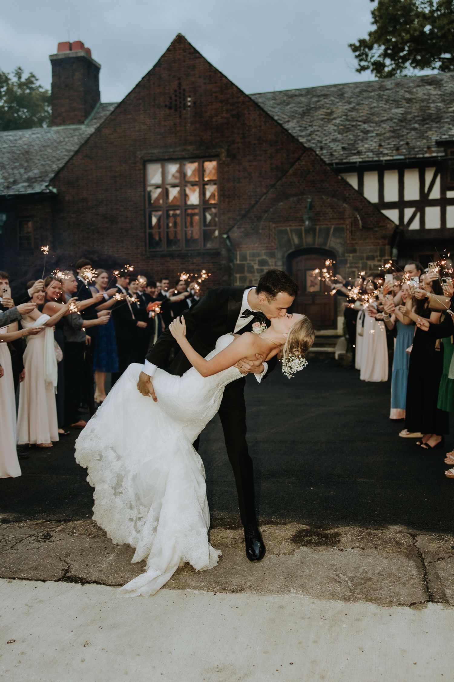Wedding party performs synchronized dance moves outdoors at dusk near a Tudor-style building.