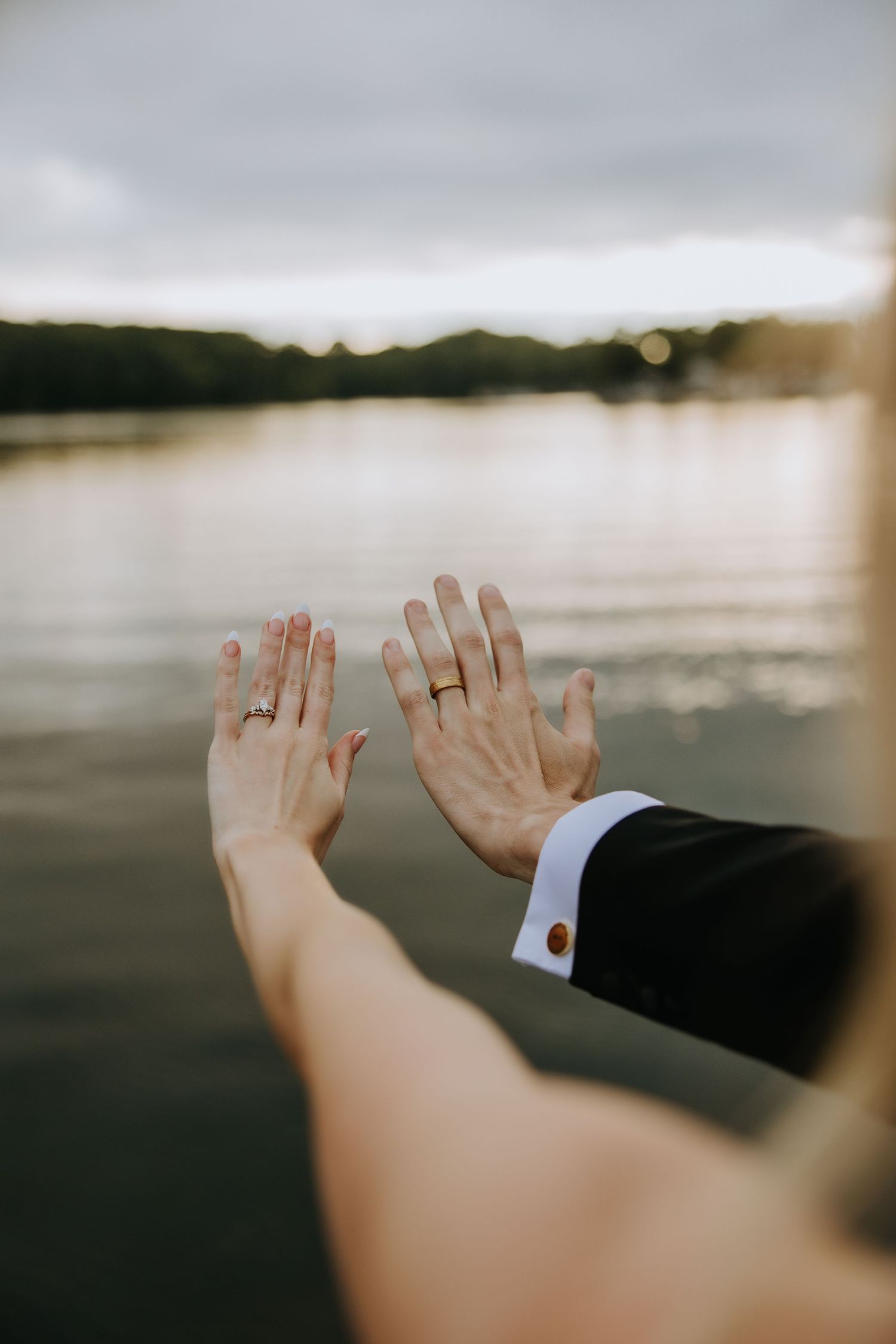 Hands with wedding rings held up against a serene lakeside backdrop at sunset.
