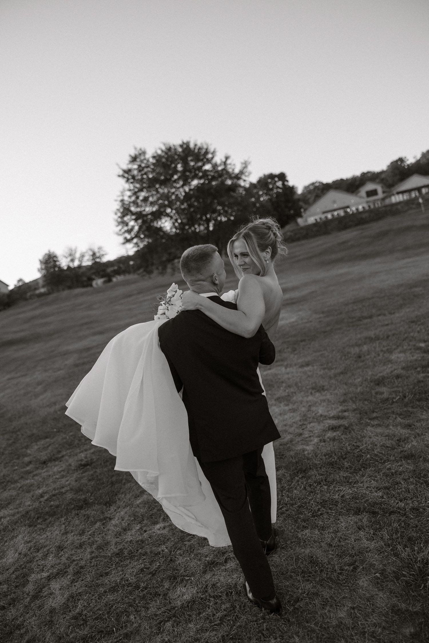 A person in black clothing dips someone wearing a flowing white dress in a dramatic dance pose on a grassy hillside.