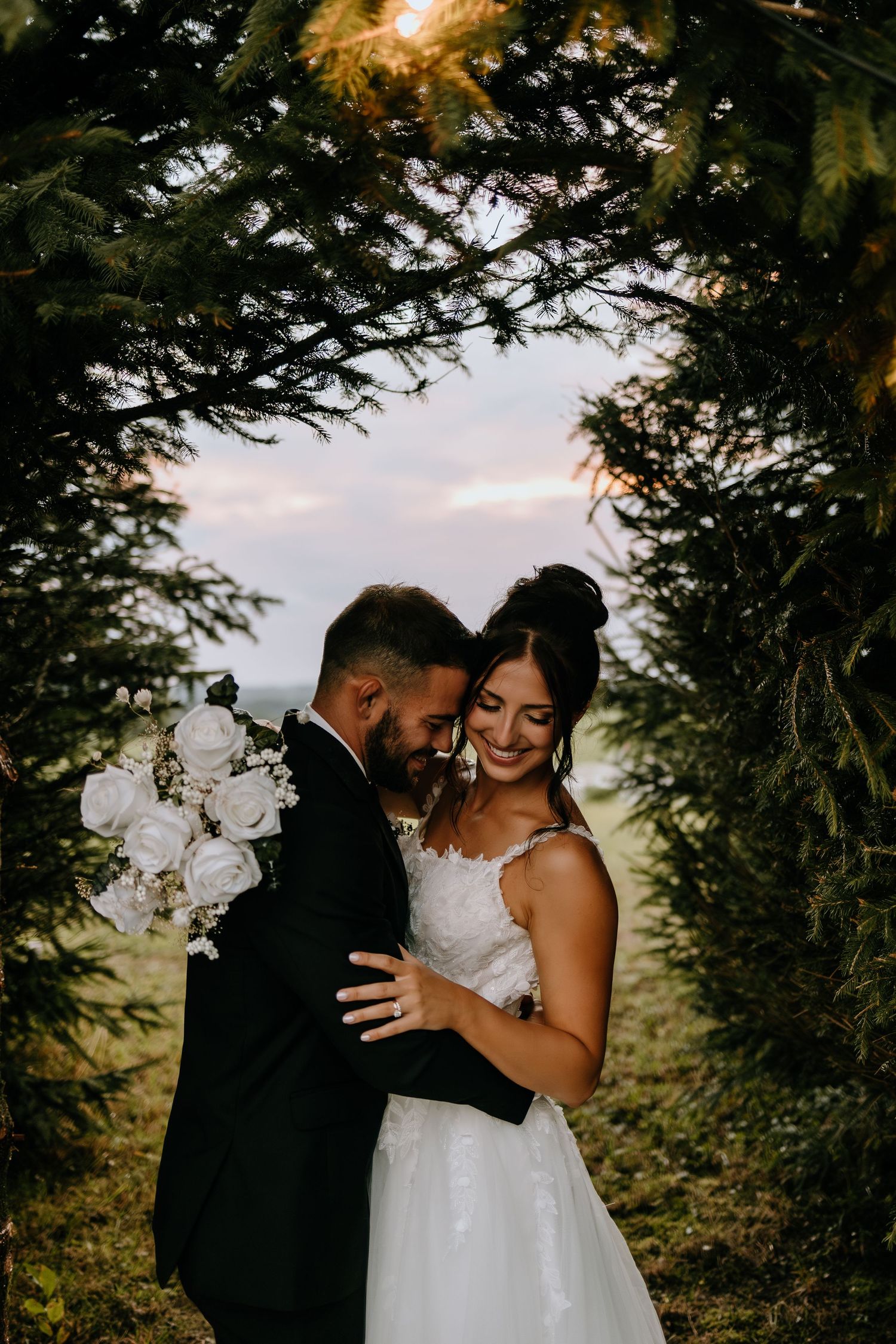 A romantic wedding couple embraces under an evergreen archway during sunset, creating an intimate woodland scene.