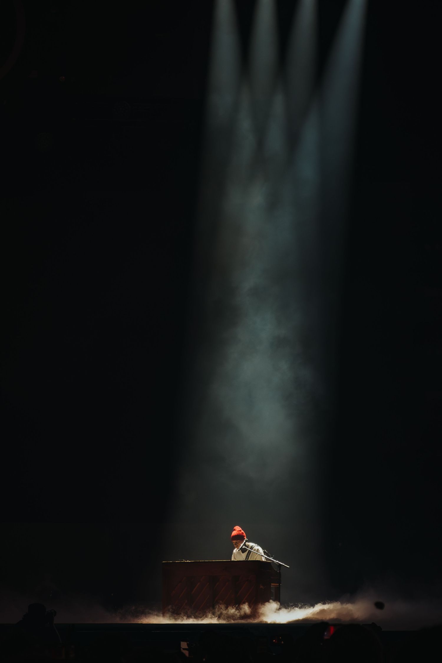 Dramatic spotlights illuminate performer at piano on darkened stage.