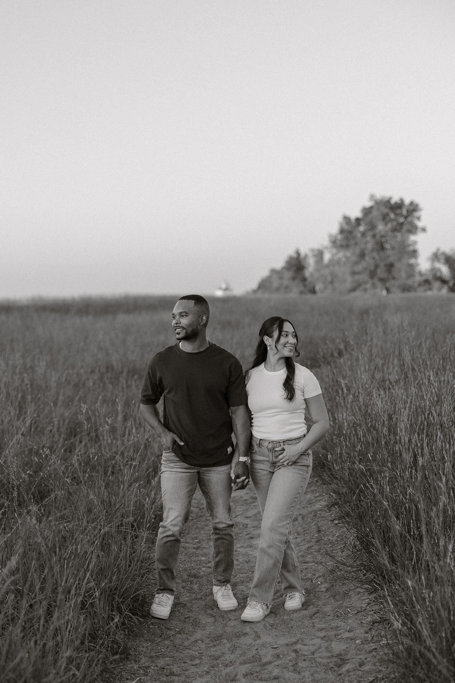 A couple stands together in a grassy field during sunset, captured in black and white.