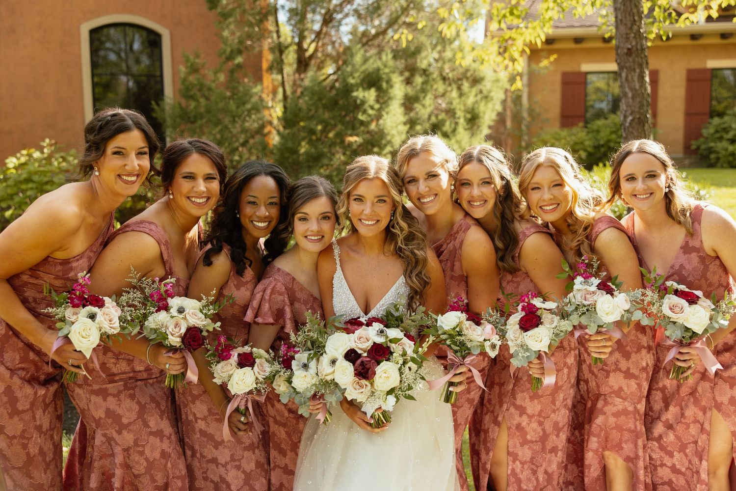 A wedding party wearing dusty rose bridesmaid dresses poses together holding white and pink floral bouquets.