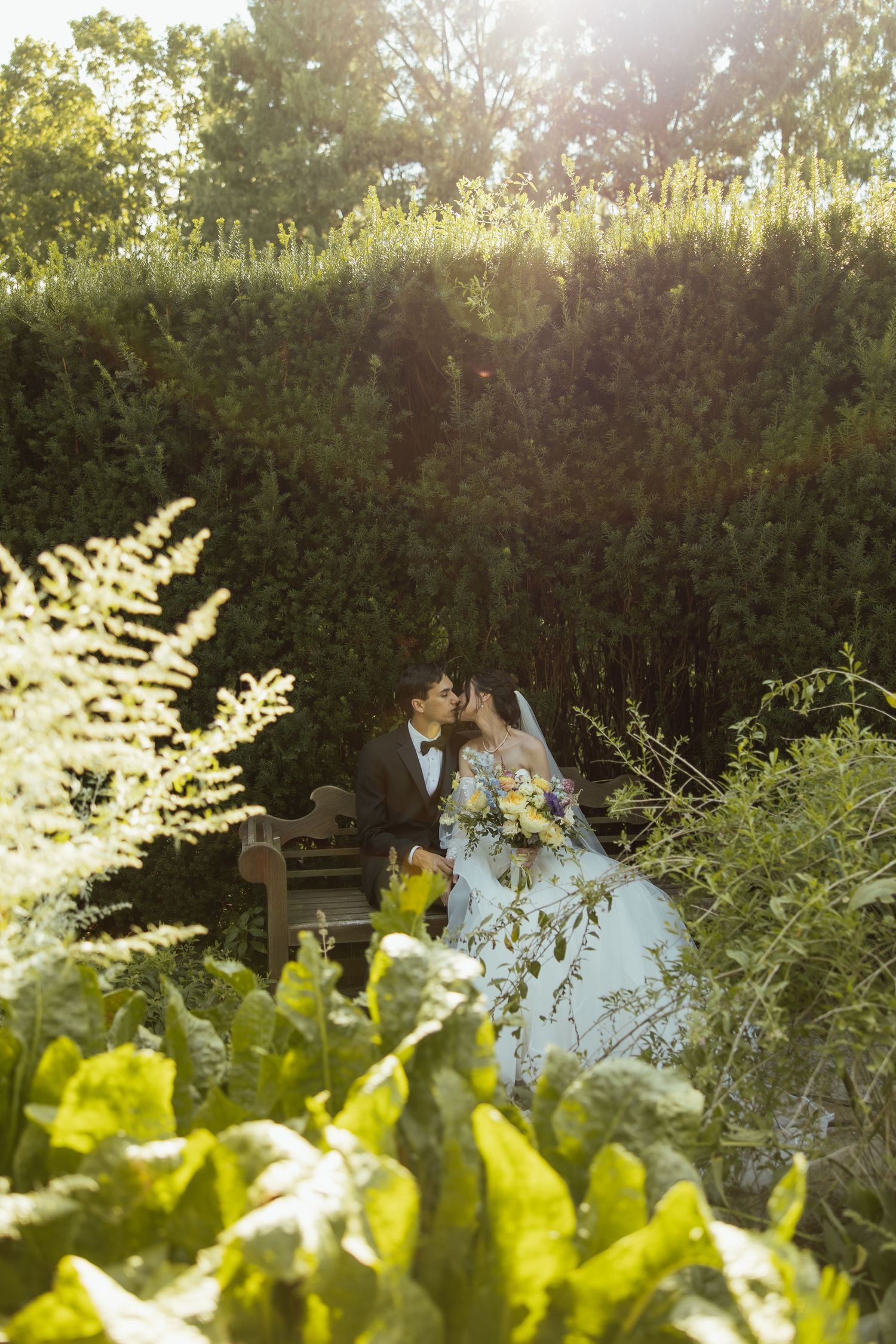 White wedding dress detail with greenery and sunlit garden hedge in the background.