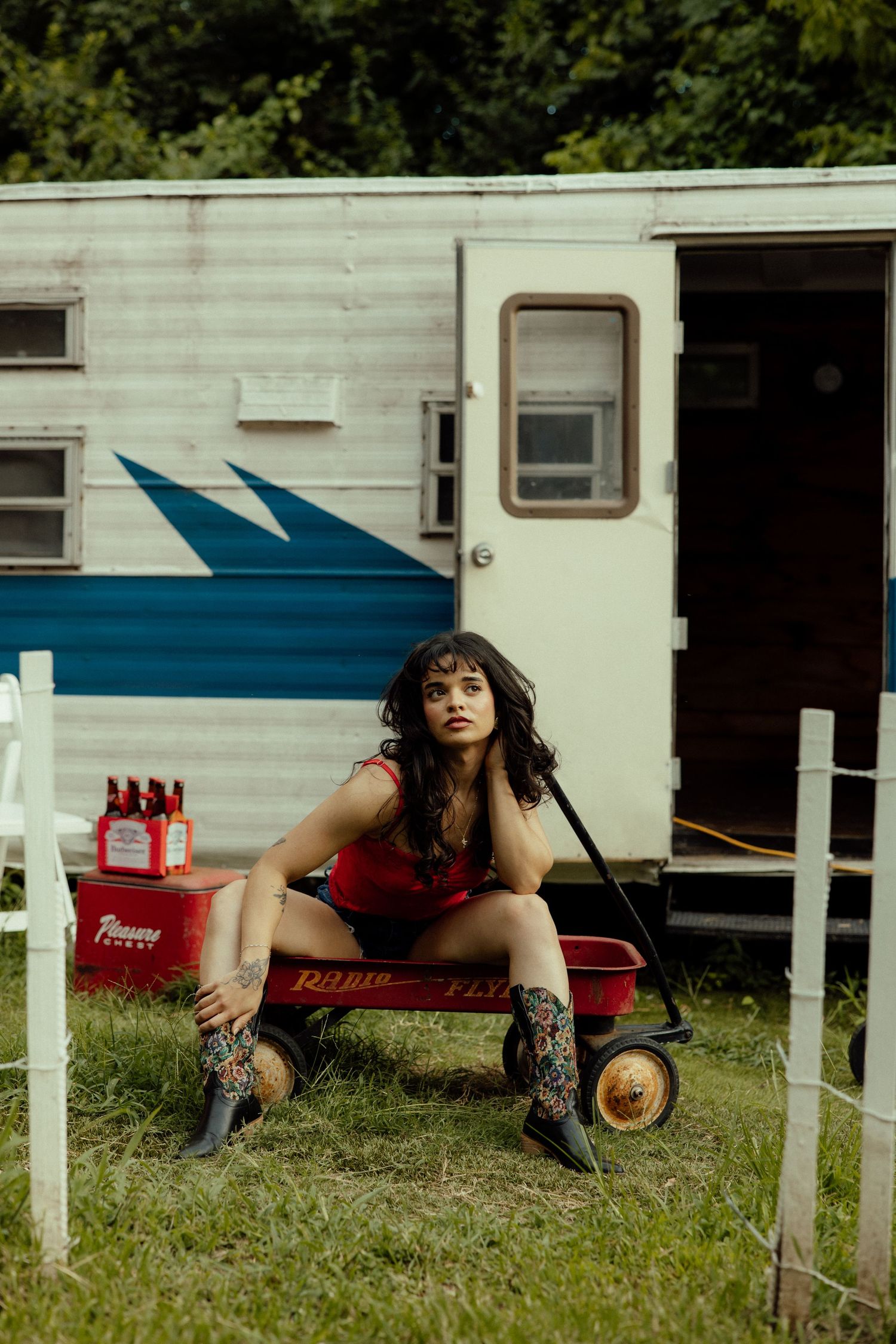 A red cooler sits on grass near a white trailer with someone lounging on it.