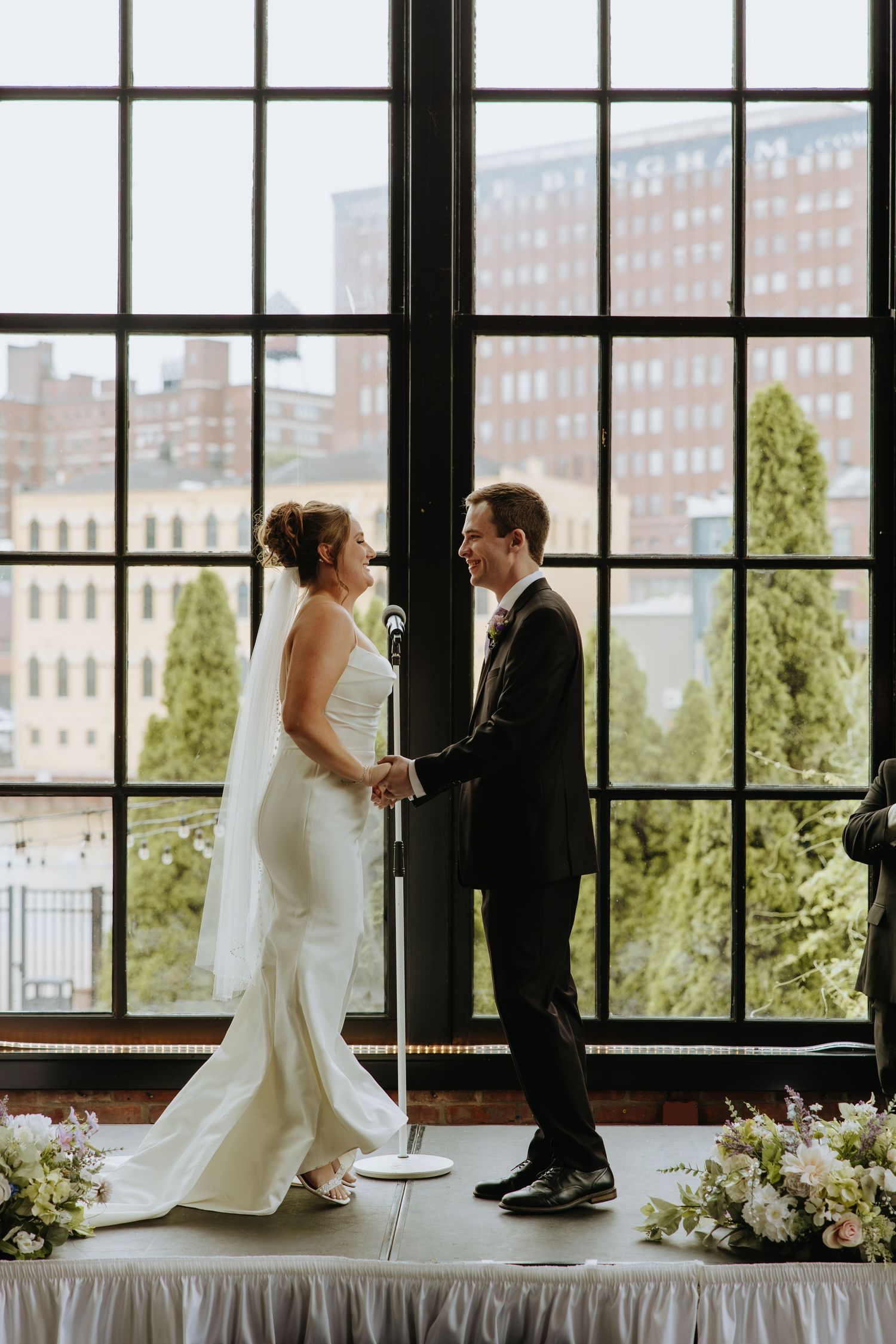 A couple shares an intimate moment during their wedding ceremony against large industrial windows with city views.