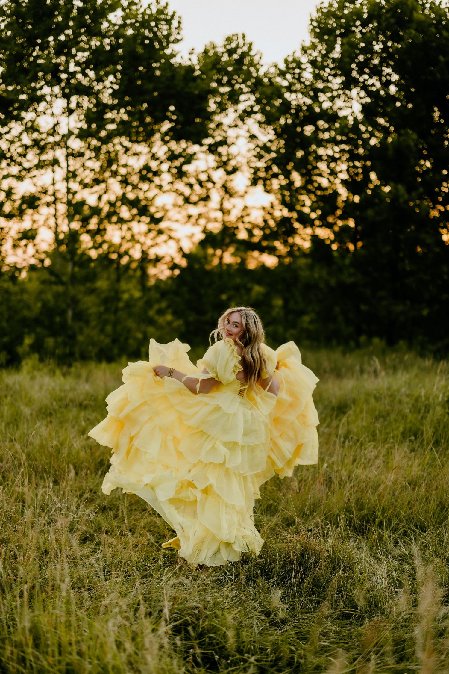 A flowing yellow dress twirls gracefully in a sunlit meadow at golden hour surrounded by trees.