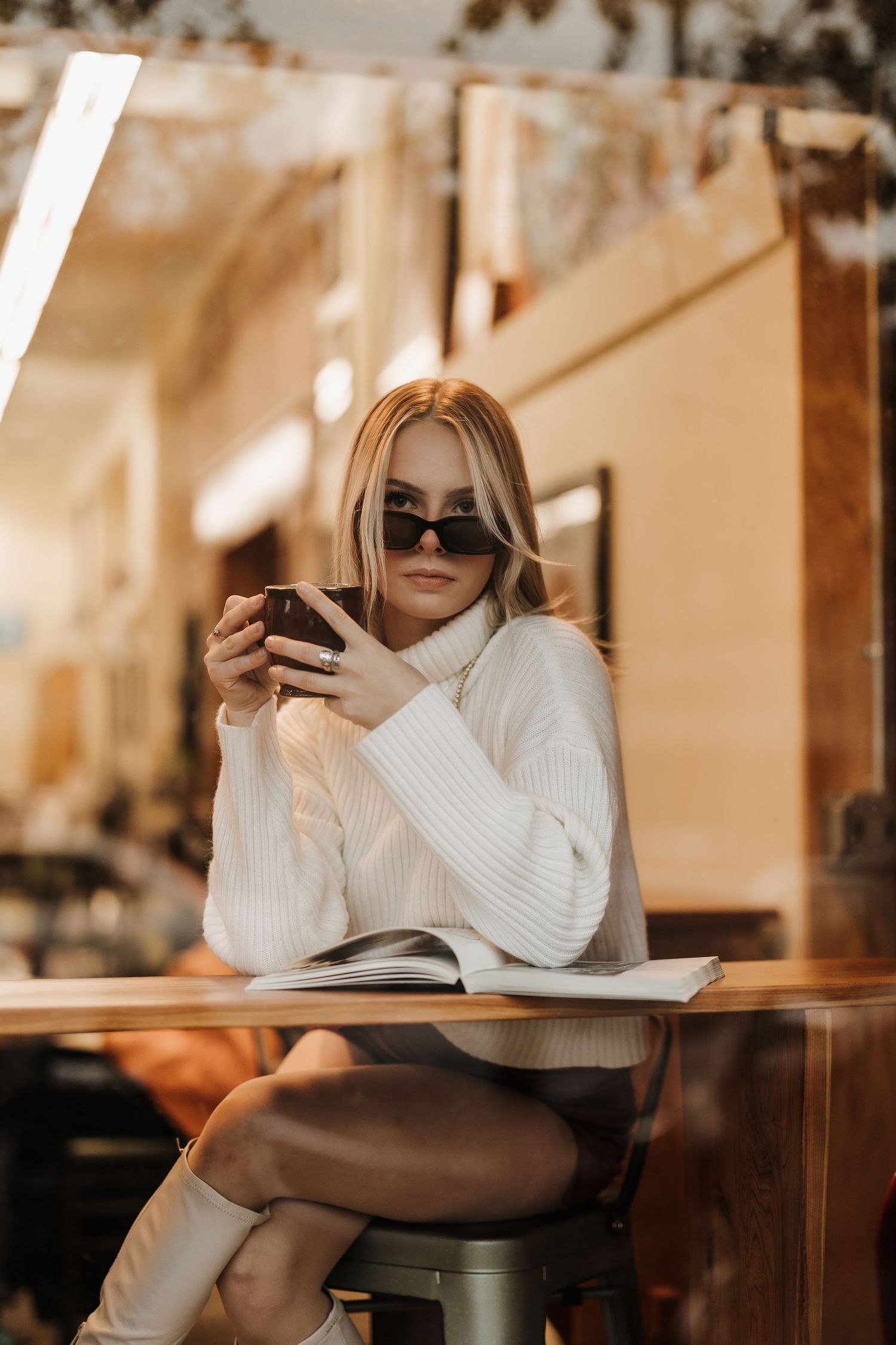 Person in white sweater and sunglasses enjoying coffee at modern cafe.
