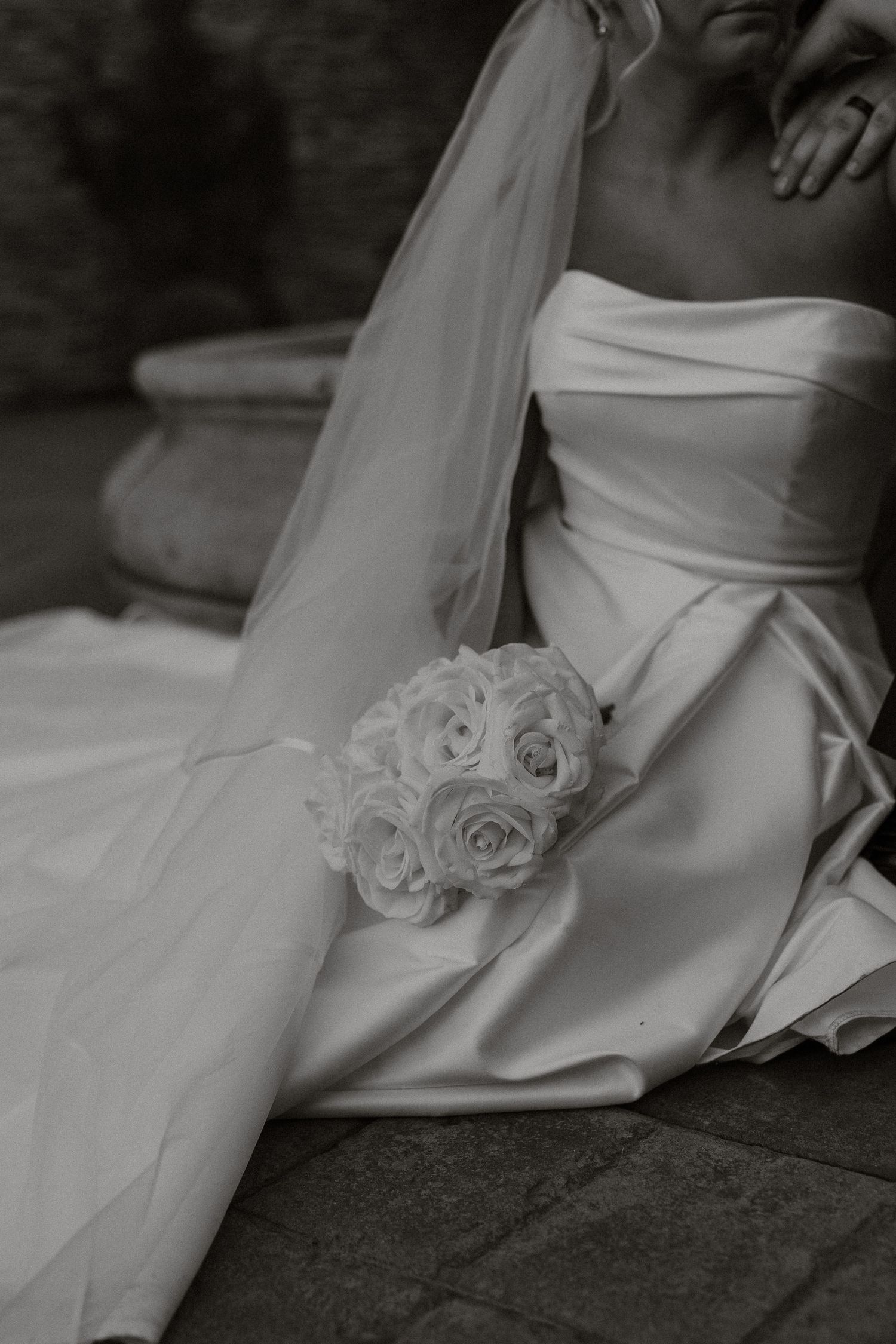 A black and white detail shot of a wedding dress with a white rose bouquet resting on its flowing fabric.