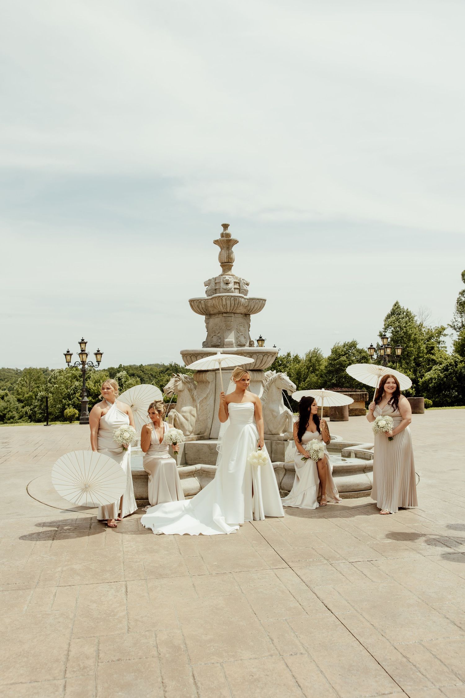 Wedding party with parasols poses around an ornate stone fountain on a sunny terrace with trees in background.