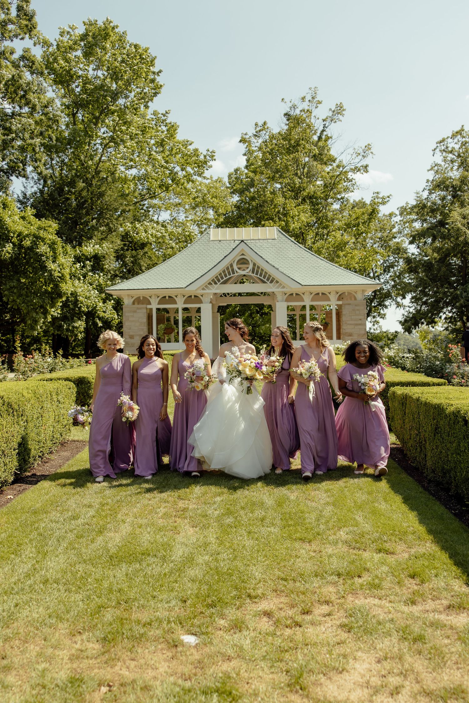 Wedding party poses in front of white gazebo with bridesmaids in purple dresses on sunny day.