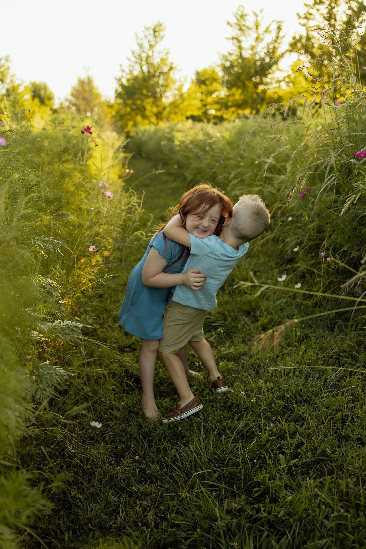 Two children play and embrace in a lush green field during golden hour as sunlight filters through the trees.