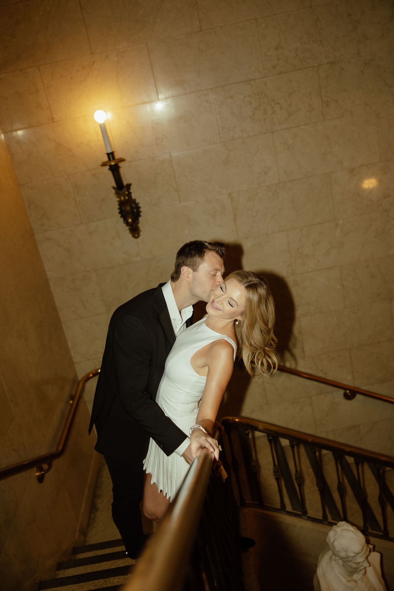 A couple shares a romantic kiss on an elegant staircase in warm lighting during what appears to be a wedding celebration.