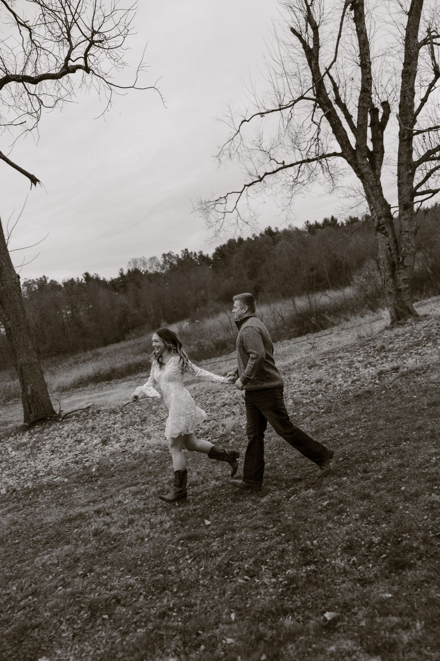 A couple walks hand in hand through a hillside meadow on an overcast day, surrounded by bare winter trees.