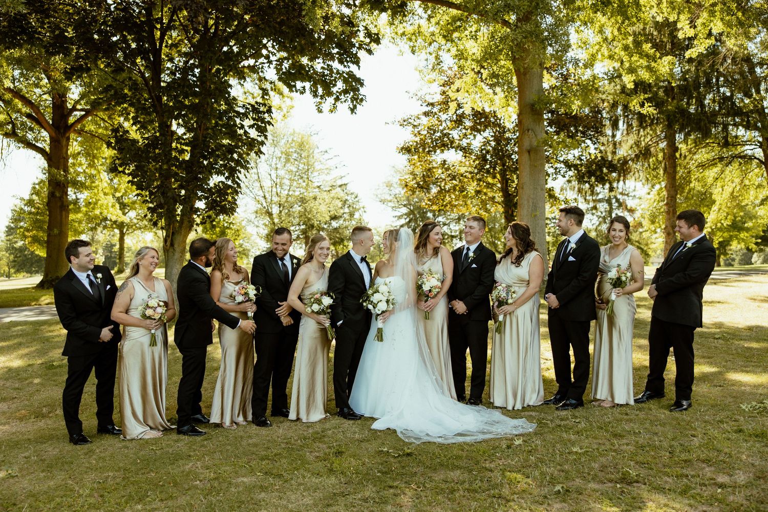 Wedding party poses for formal group photos under sunlit trees during golden hour.