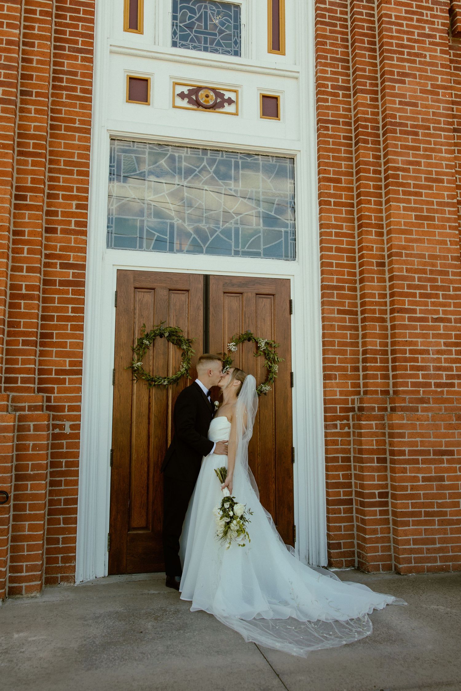 Elegant bride in flowing white gown stands against red brick church entrance with decorative greenery and wooden doors.