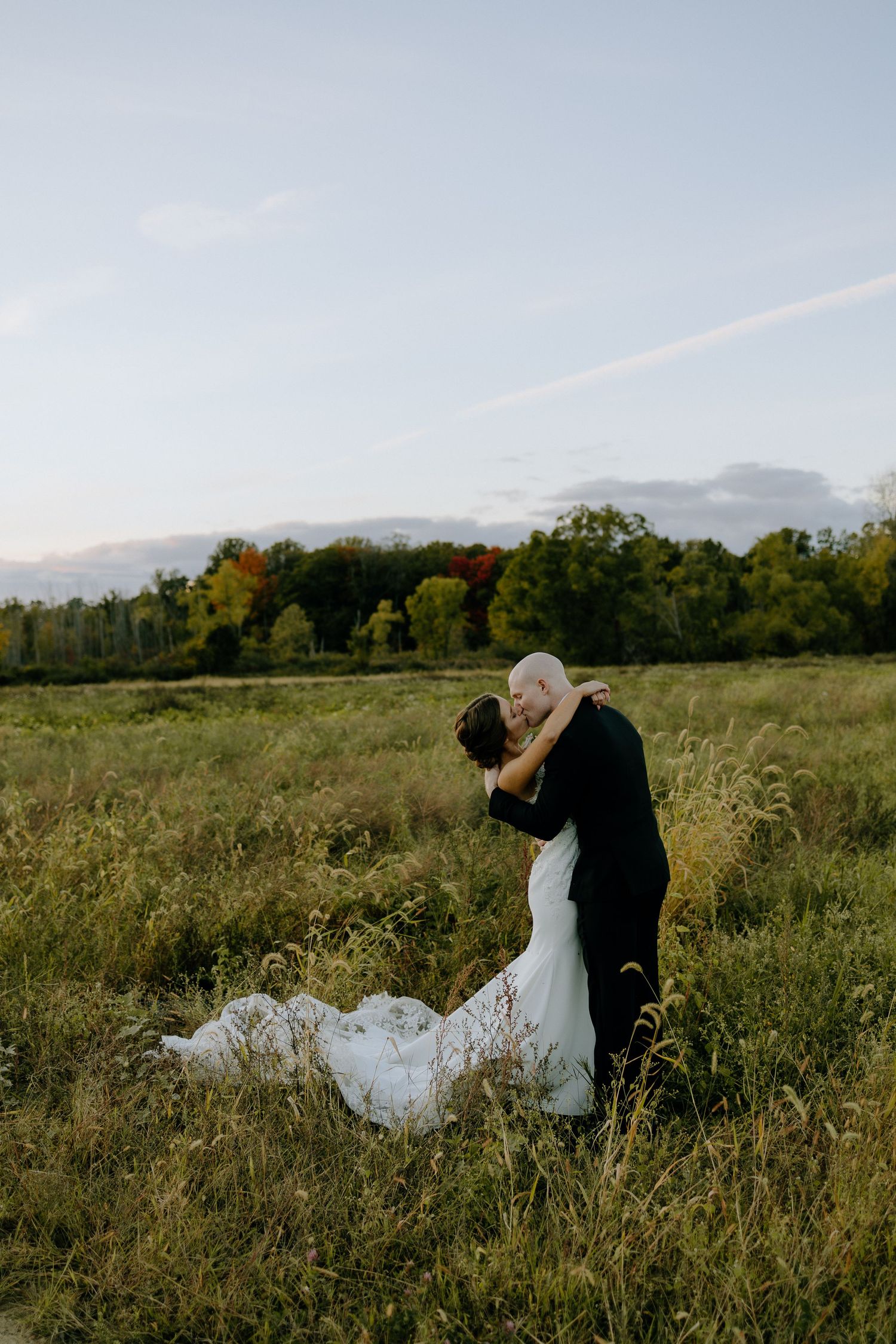Two people embrace in a scenic meadow with mountains and trees in the background during golden hour.