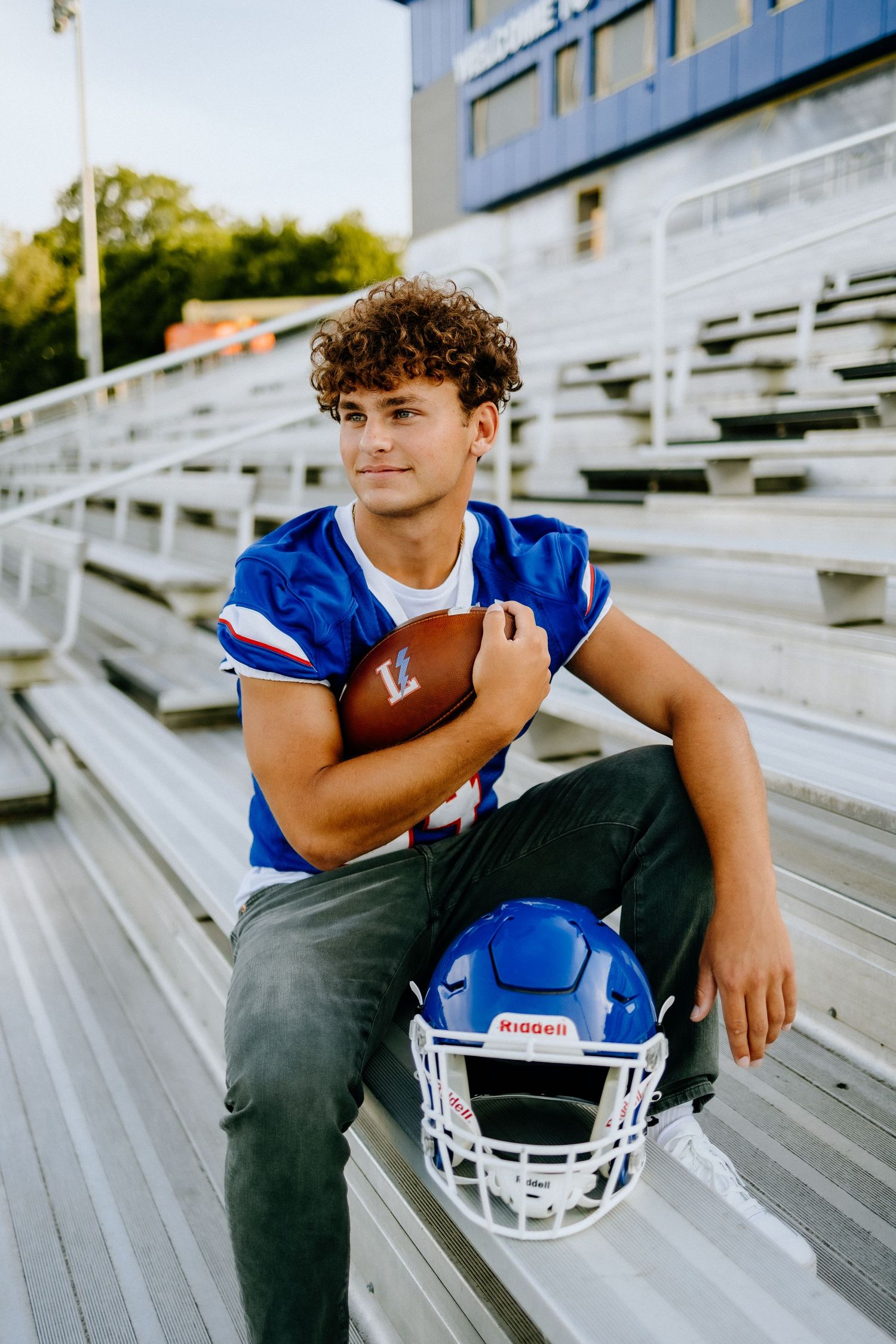 Football player in blue jersey poses with ball and helmet on stadium bleachers.
