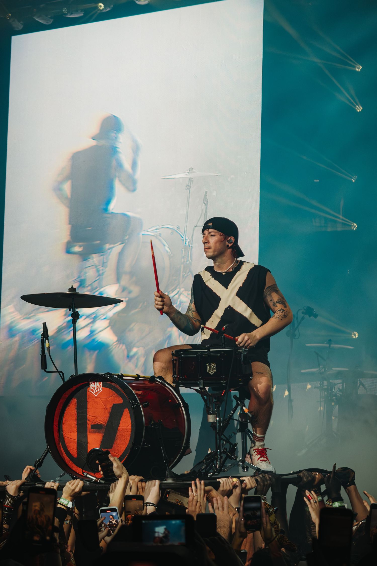 Drummer performs elevated on stage with dramatic lighting and projection screen in background.