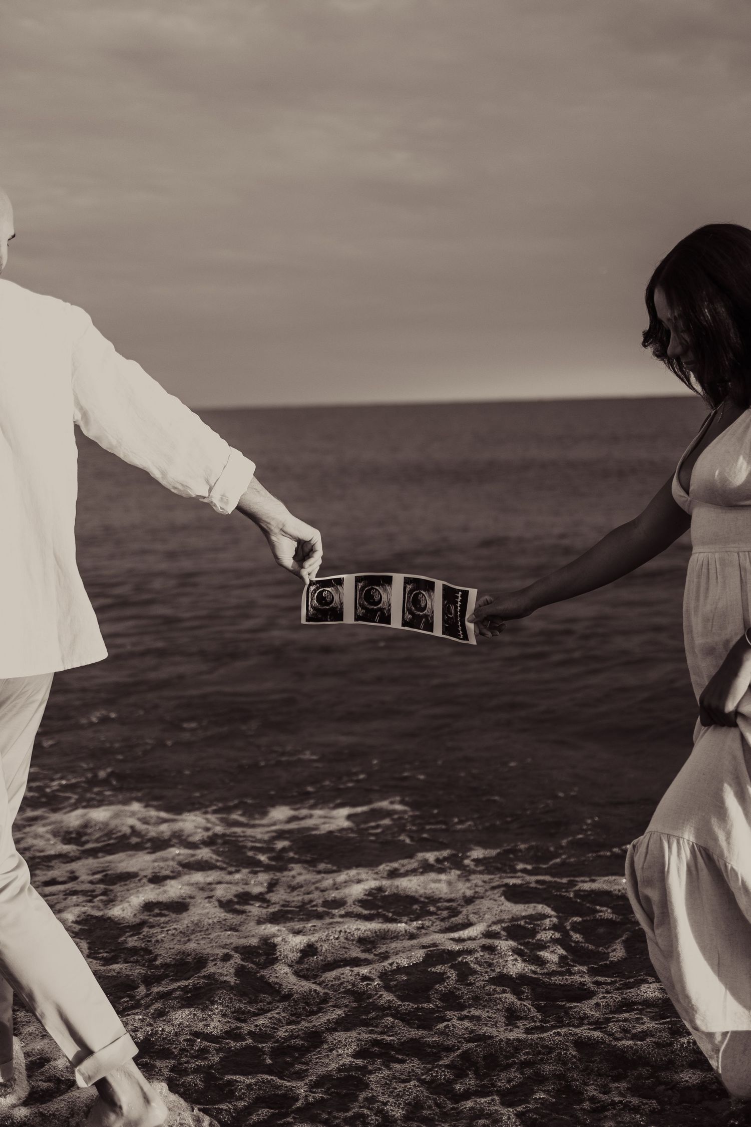 Black and white beach photo of a couple holding ultrasound photos by the ocean.
