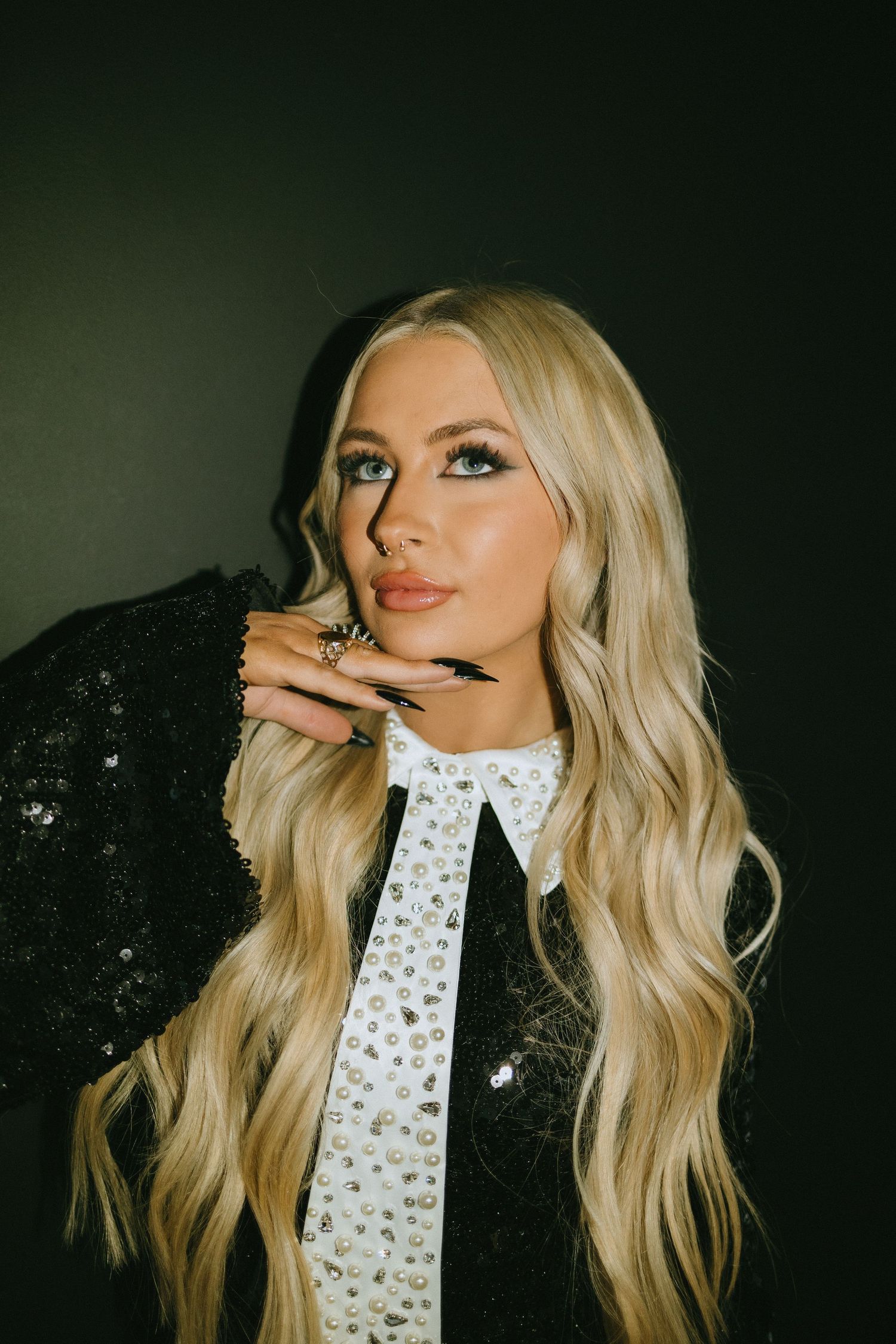 Fashion model wearing a black sequin outfit with white crystal tie and long blonde wavy hair against dark background.