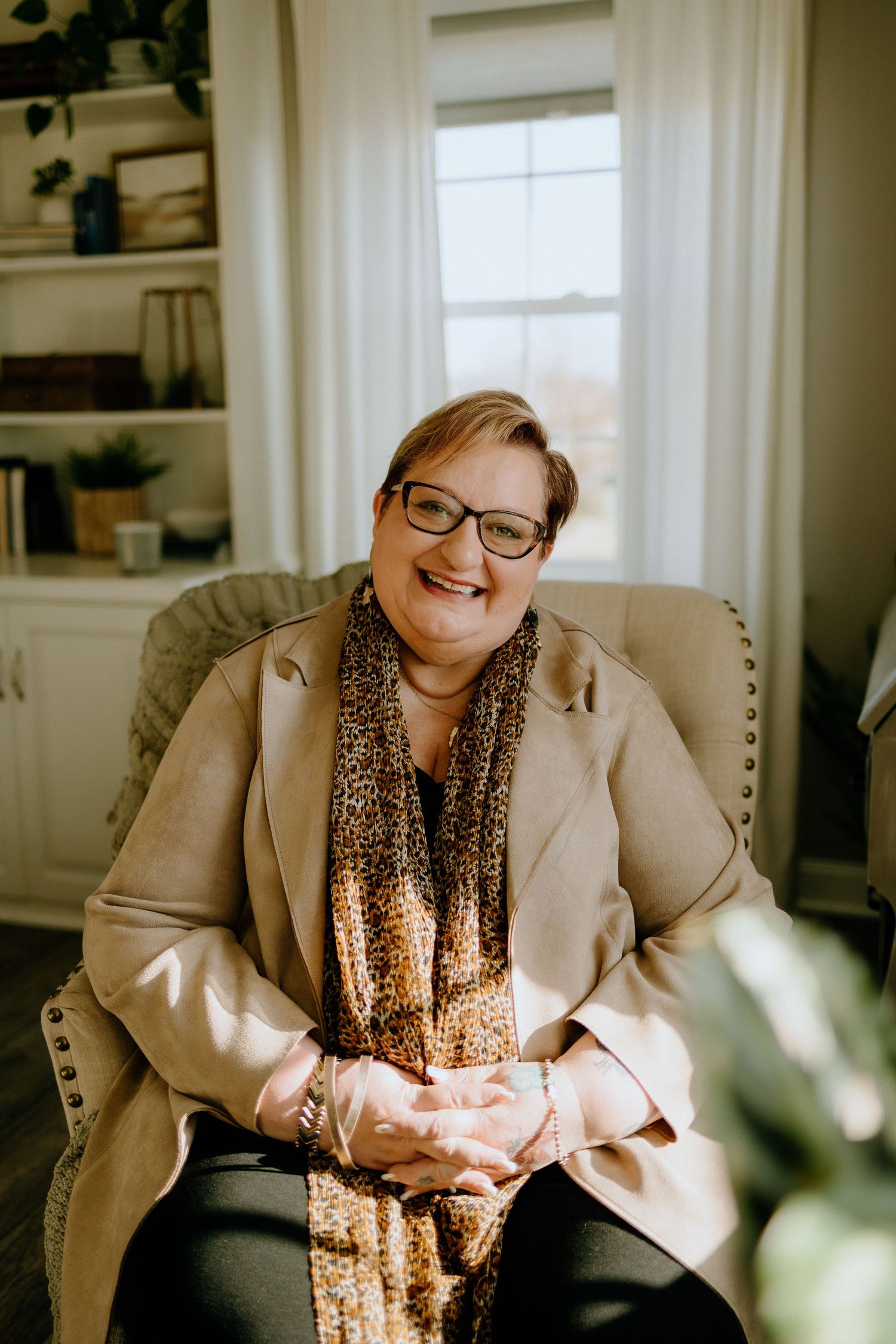 Professional wearing a beige blazer and leopard print scarf sits smiling in a home office setting.