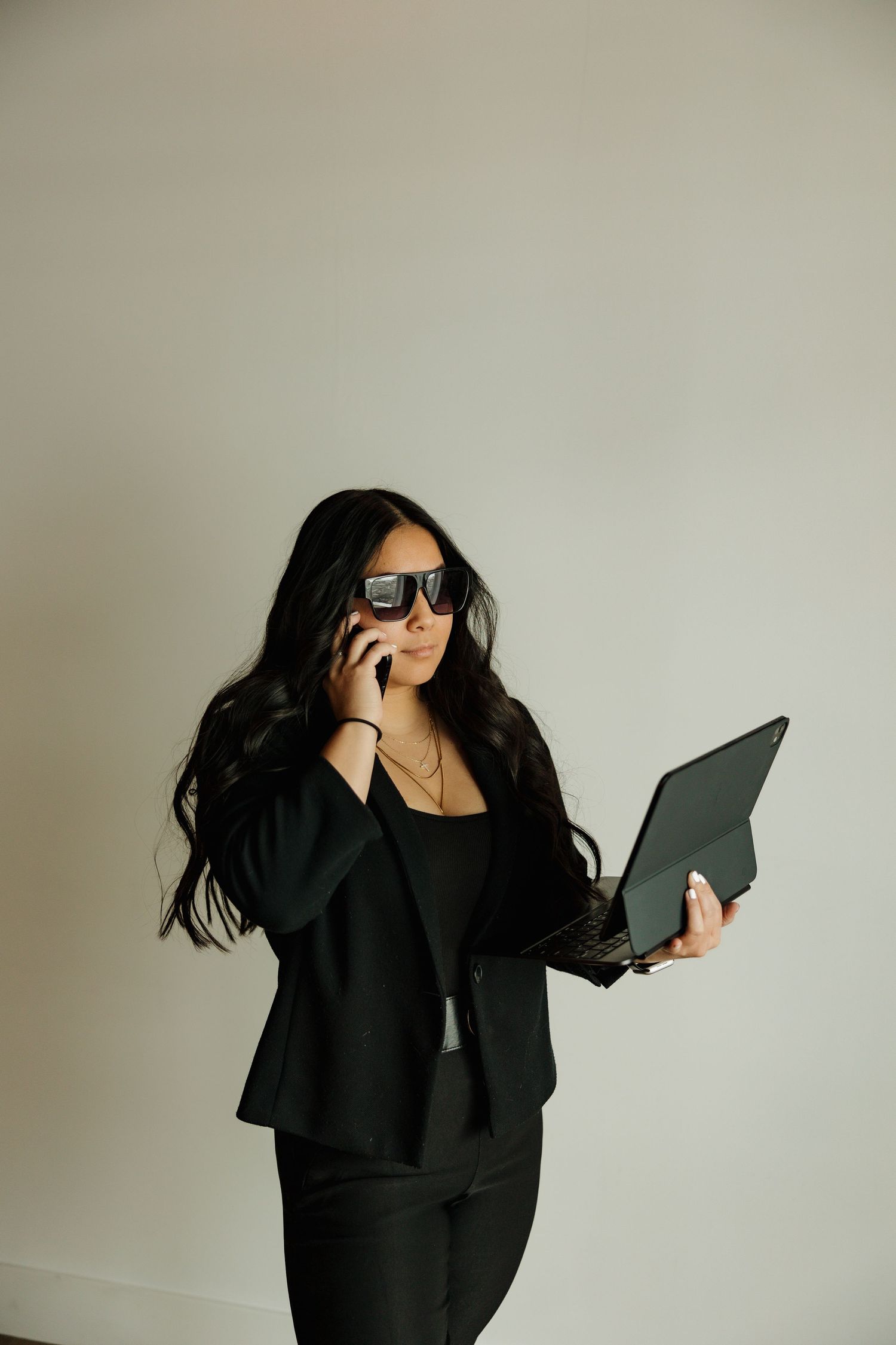 Business professional in black suit talking on phone while holding tablet and wearing sunglasses.
