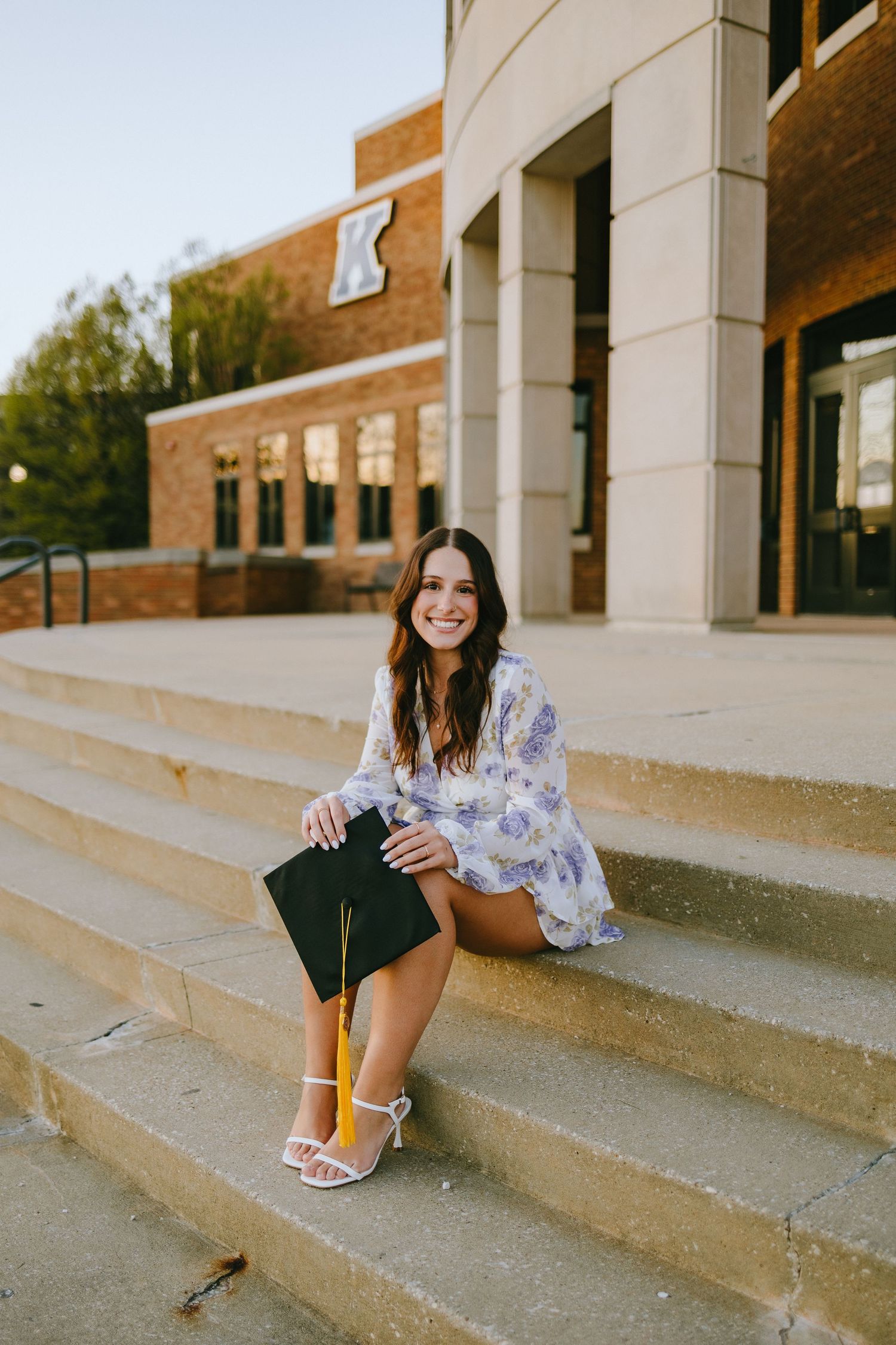 A graduate in a floral dress sits on concrete steps in front of a university building with a K logo.