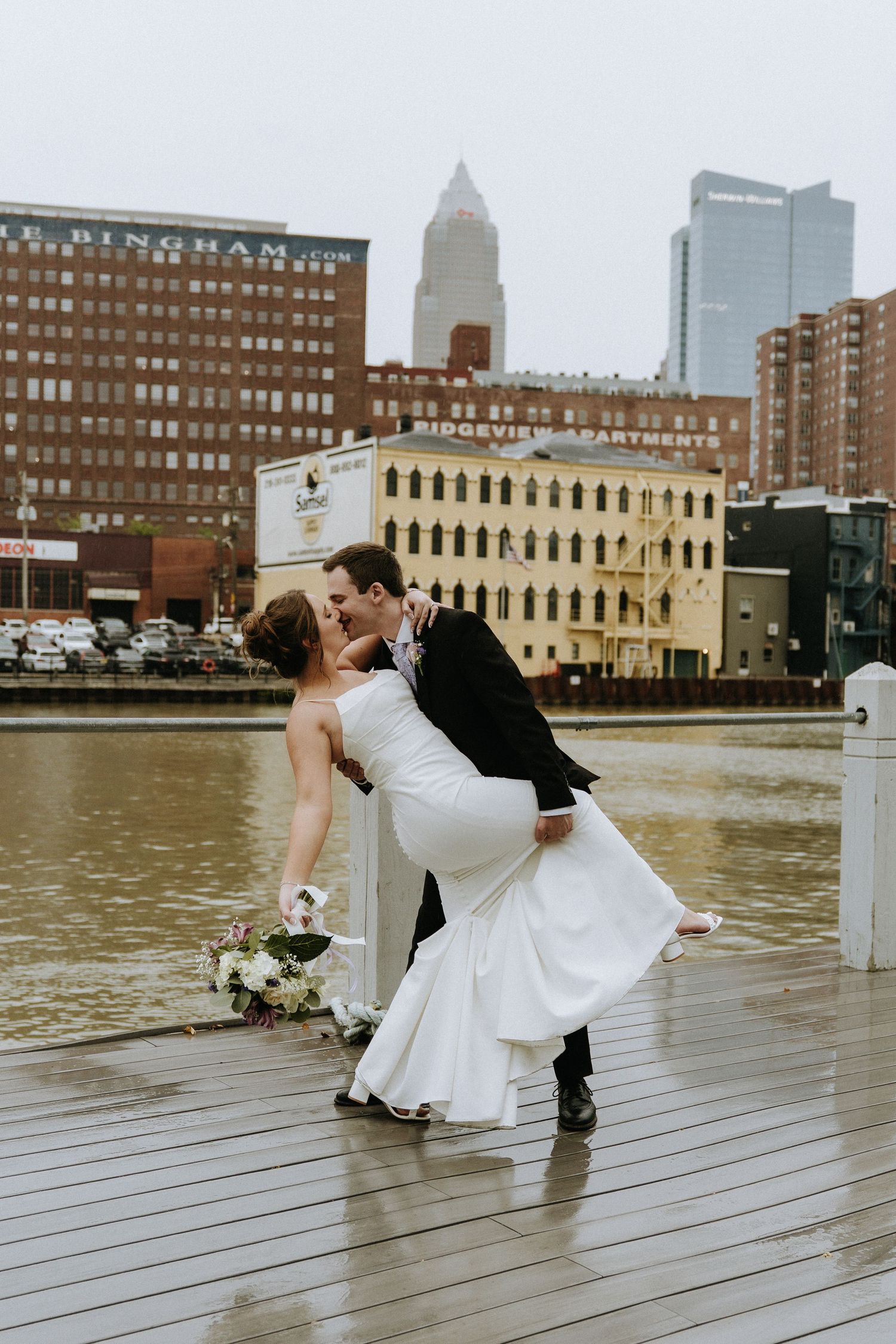 Dramatic dip pose during wedding photoshoot on wooden pier with urban waterfront backdrop.