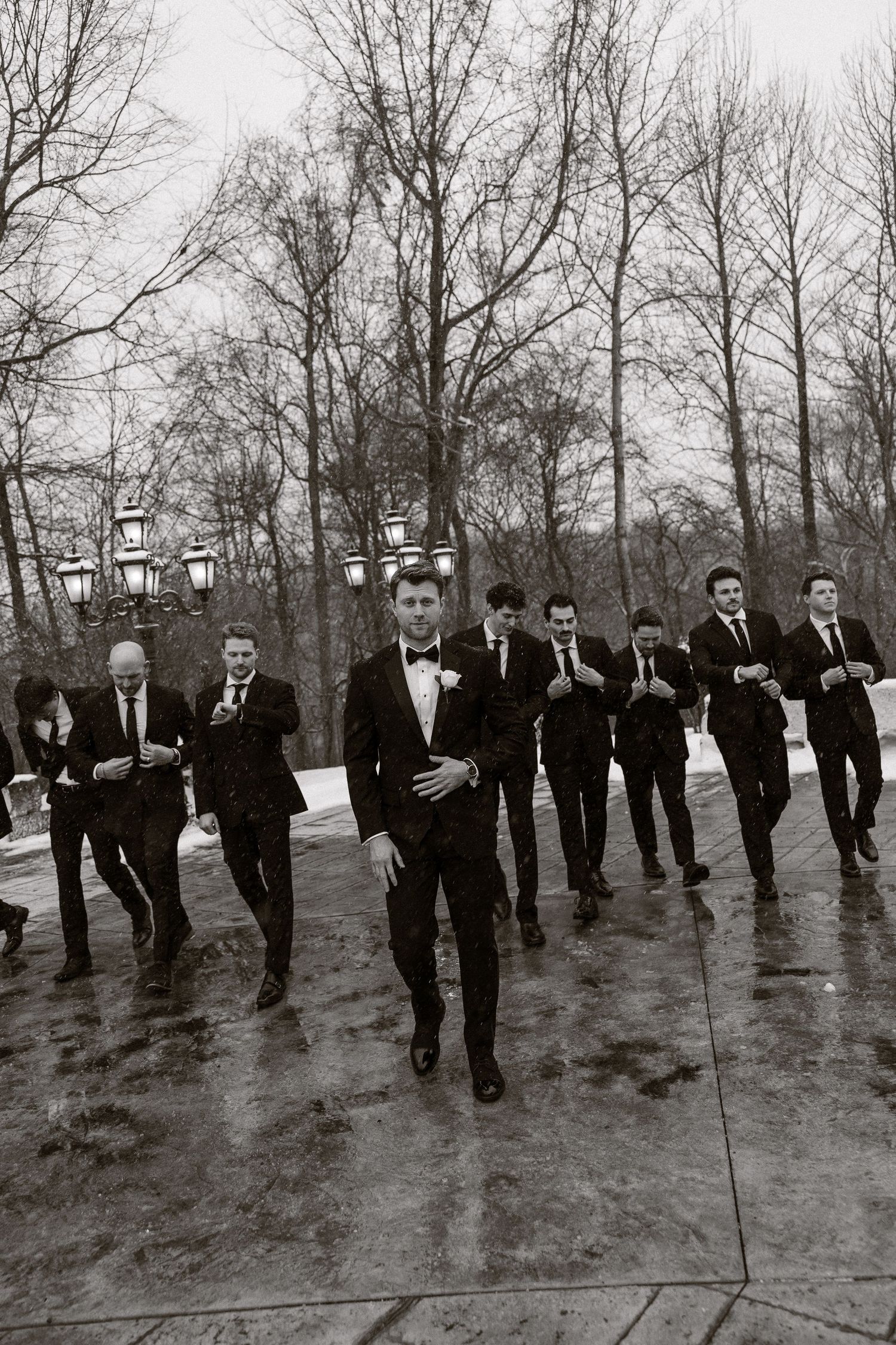 A line of groomsmen in black suits pose outdoors in a winter landscape with bare trees in the background.