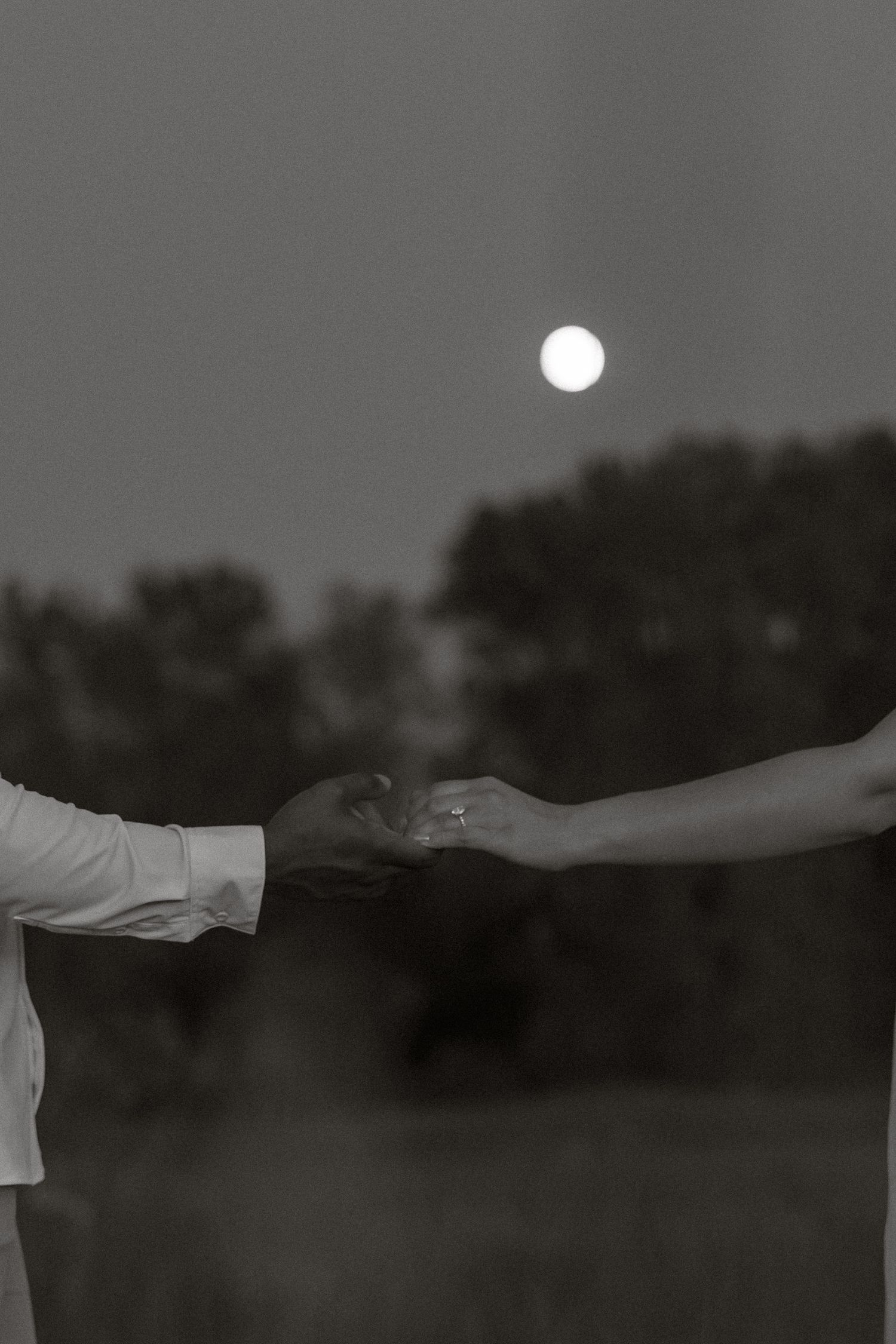Two hands reaching towards each other against a moonlit sky in a romantic black and white photograph.