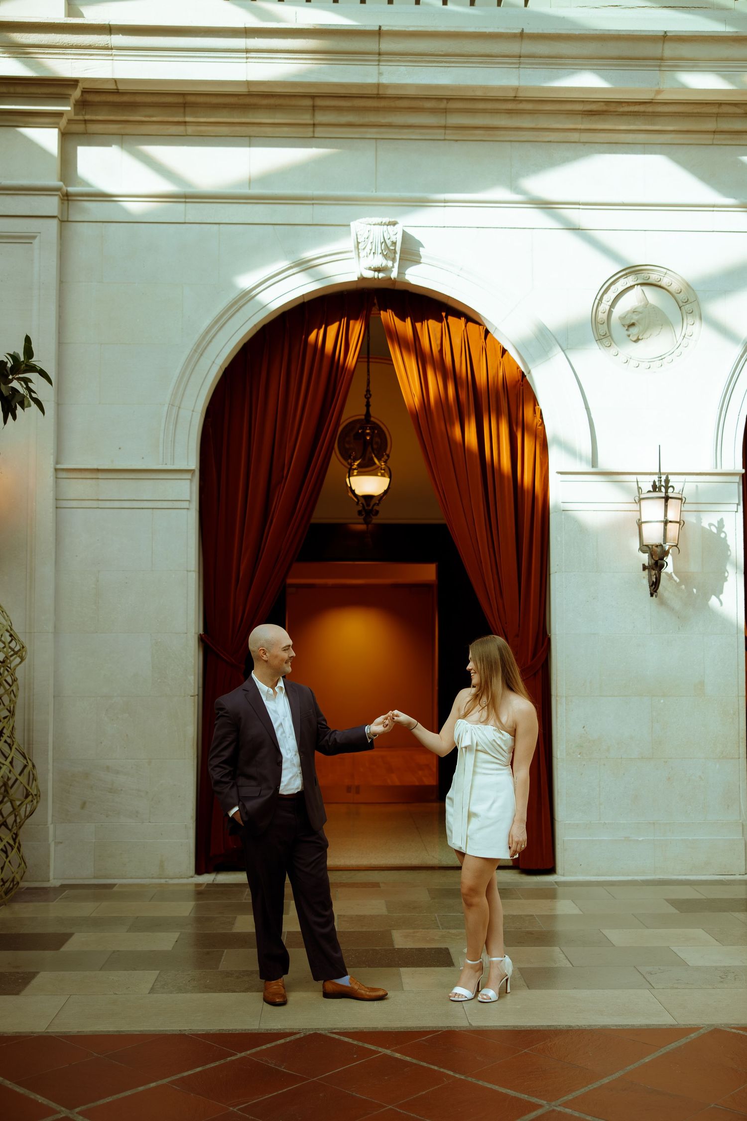 Two people stand in conversation by an elegant arched doorway with red velvet curtains and terra cotta flooring.