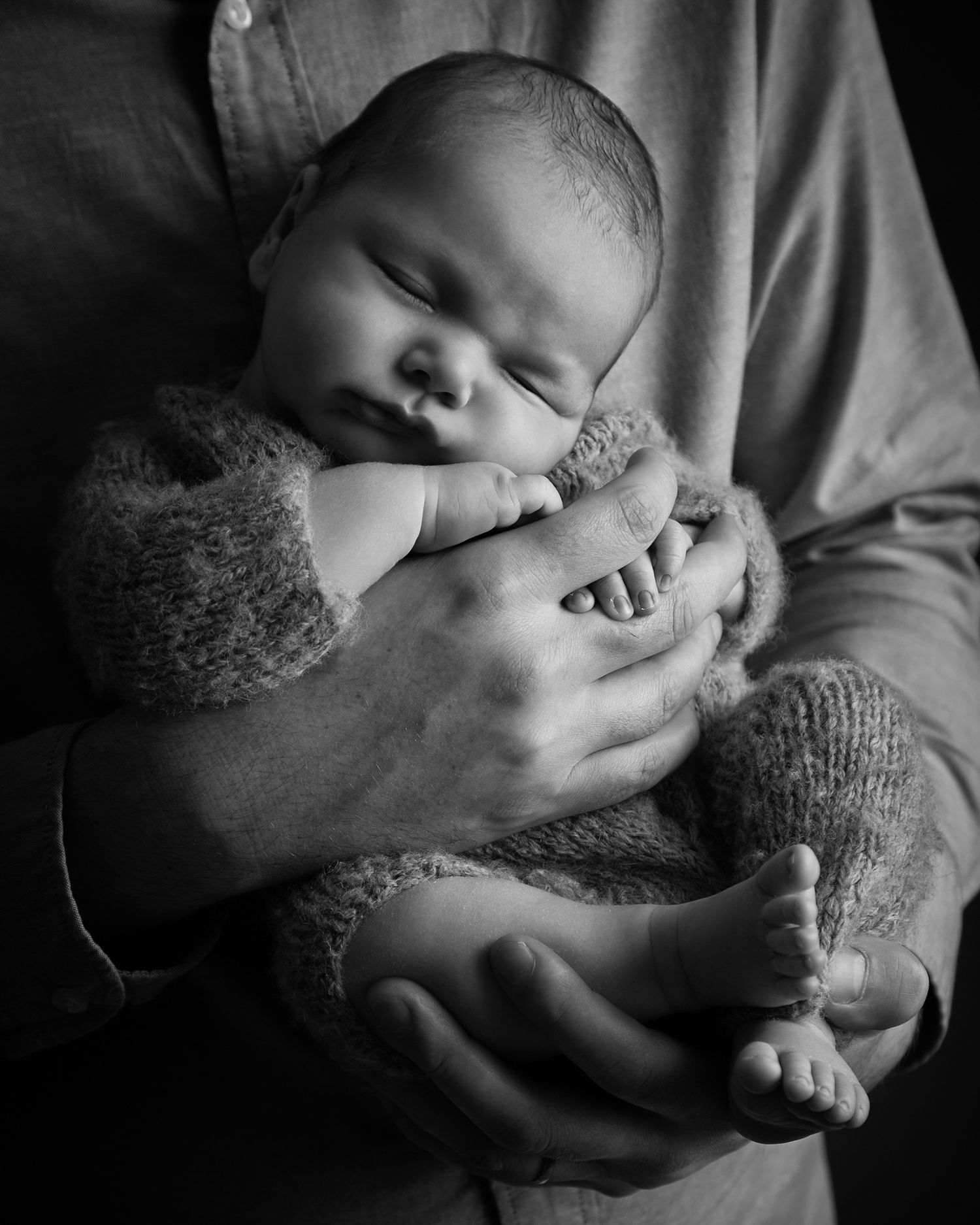 newborn baby in a soft knitted outfit held in their fathers hands in black and white