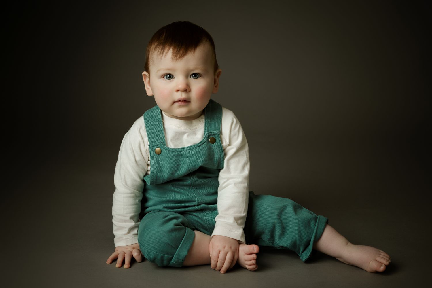 Toddler in green overalls sitting on a grey background.