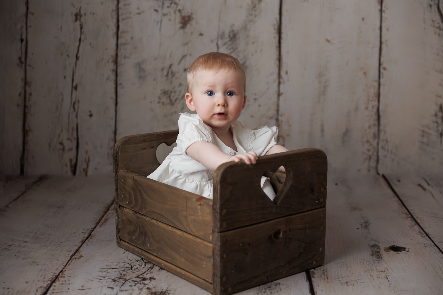 Baby in a white dress sitting inside a wooden box against a rustic wooden backdrop.